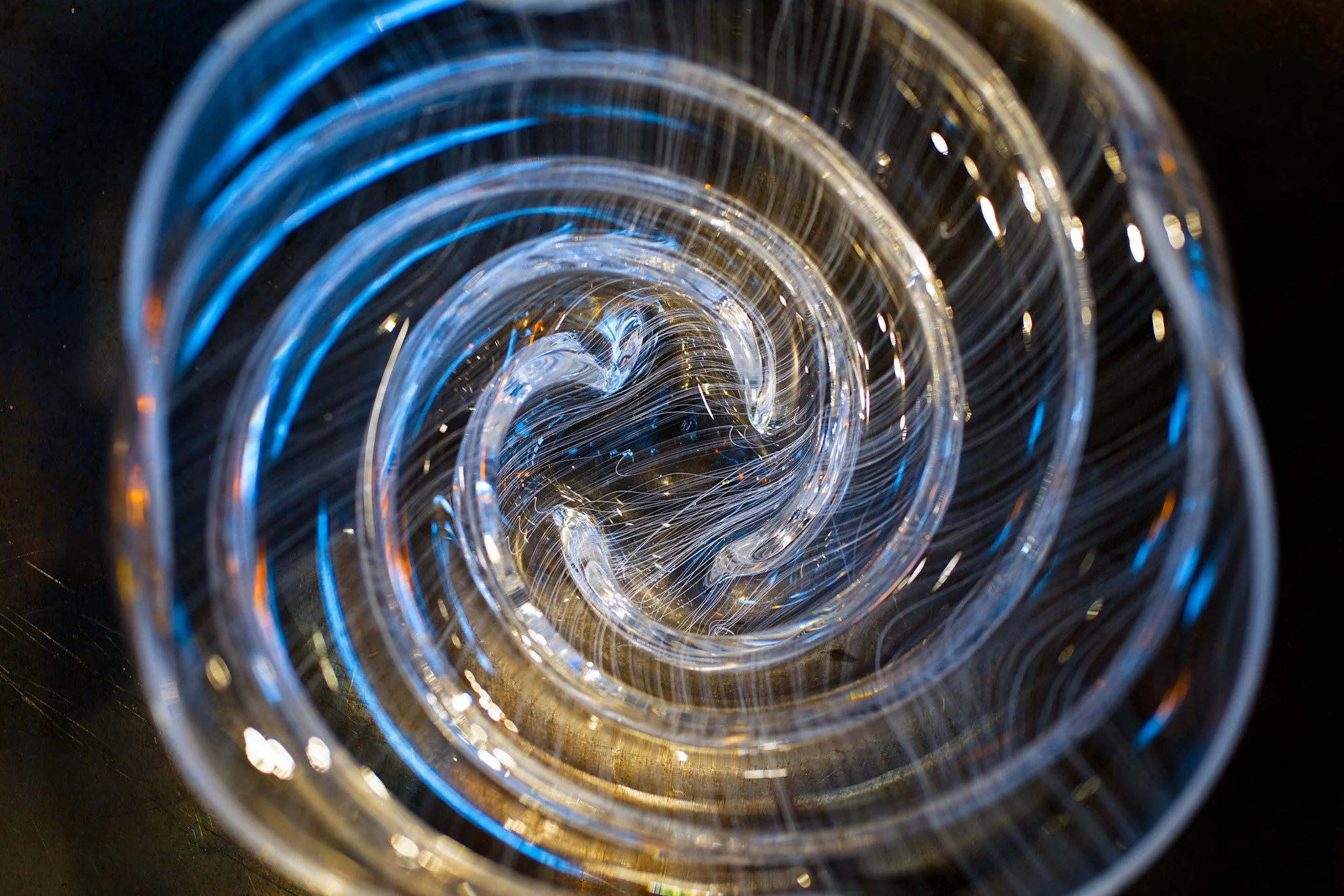 Close-up of a coiled glass object illuminated with blue and yellow light, seen from above against a dark background.