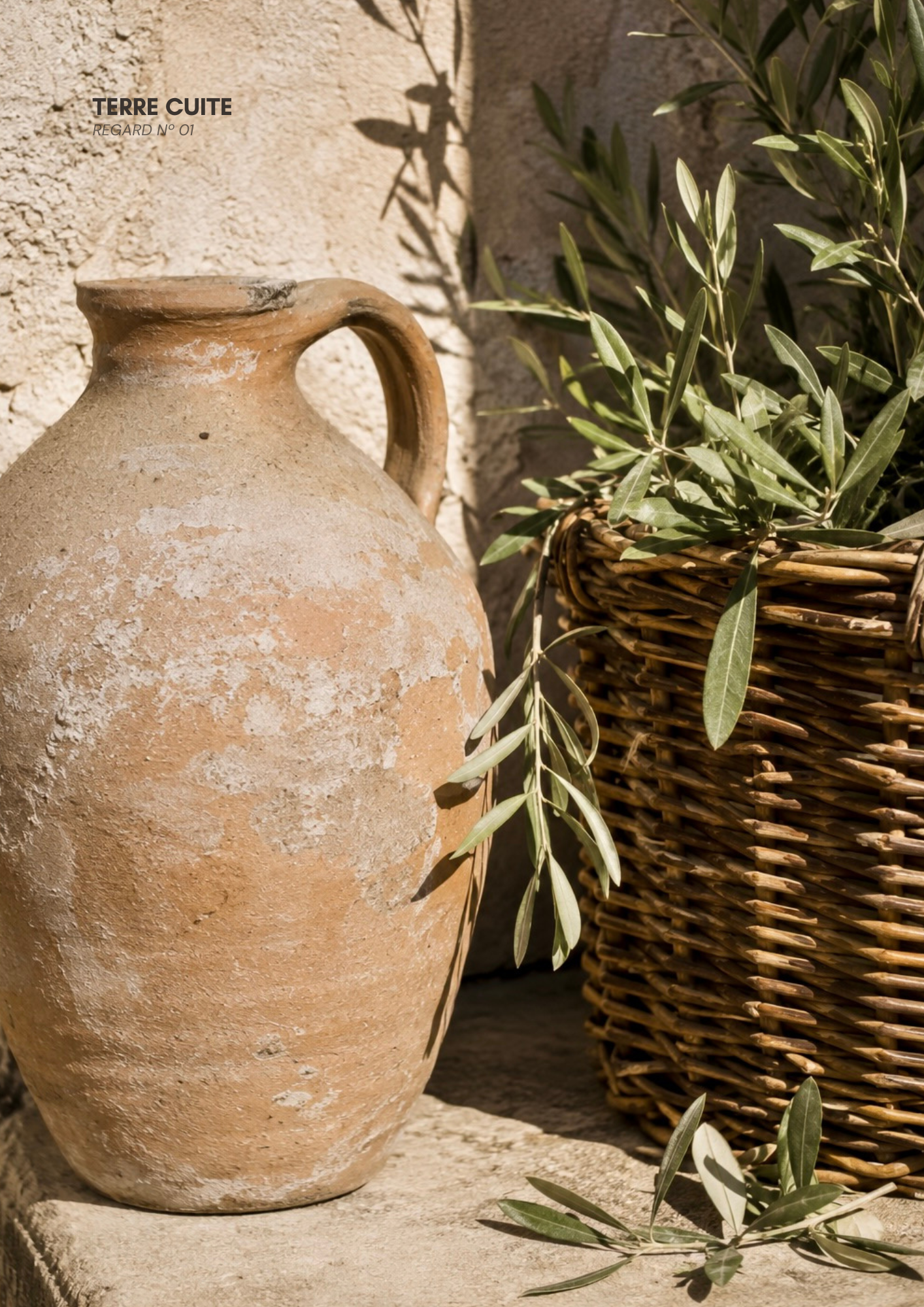 Un pot en terre cuite avec une branche d'olives à côté et un panier en osier dans un décor extérieur.