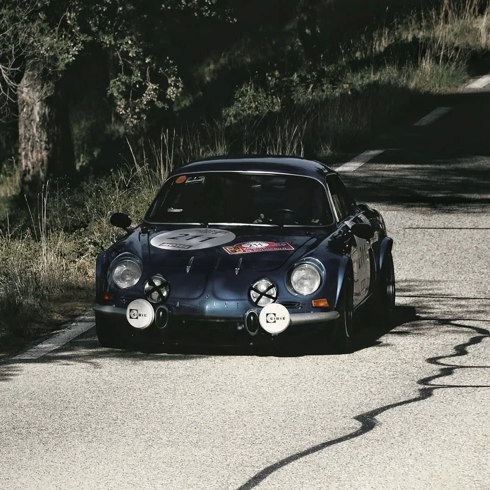 Une voiture de course classique, probablement une Porsche 911, roulant sur une route de campagne de nuit, équipée de phares supplémentaires.
