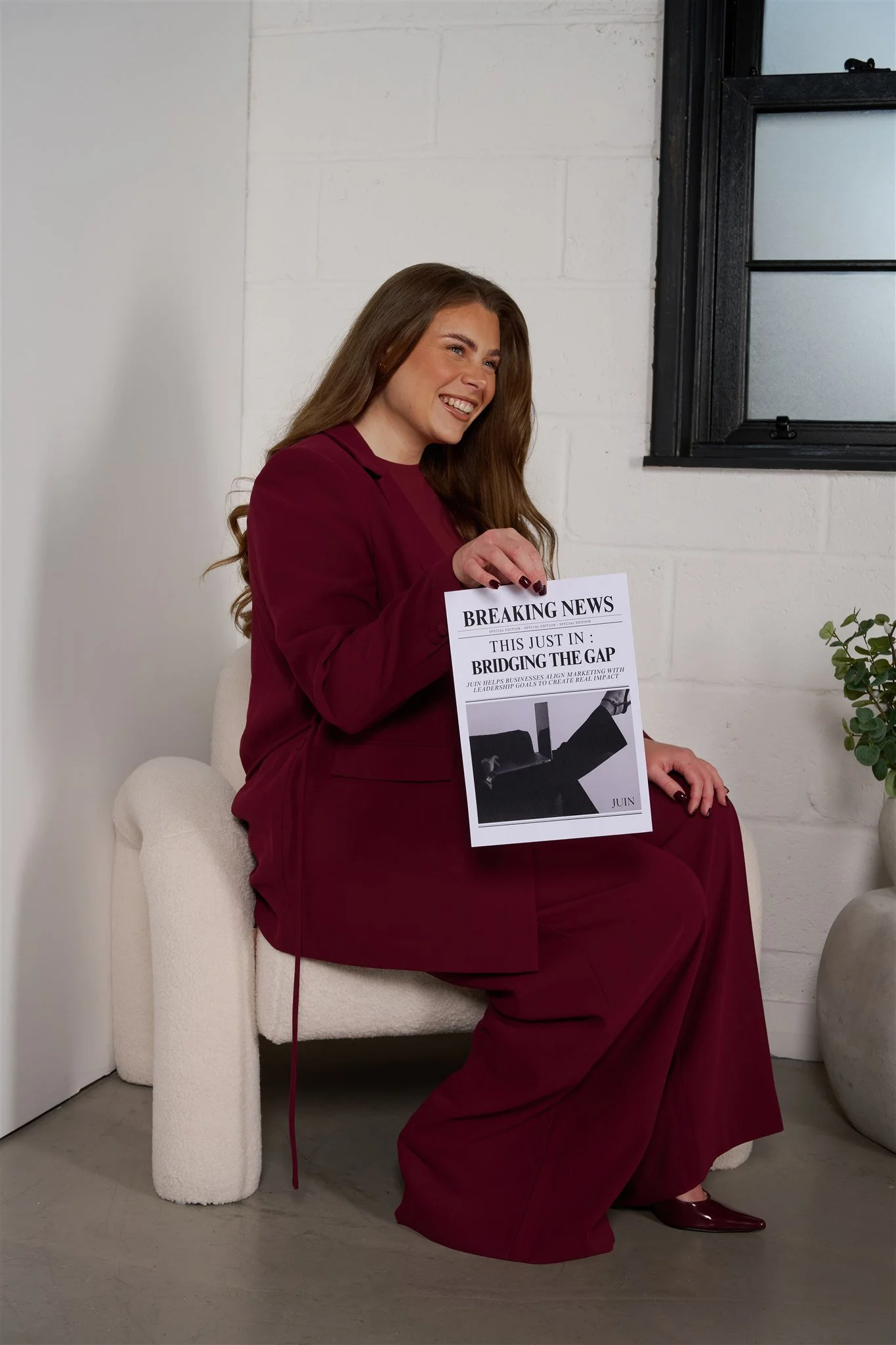 A woman sitting on a cream-colored armchair, holding a newspaper titled 'Breaking News' with headlines about bridging the gap. She is smiling and wearing a maroon suit and matching wide-leg pants. There is a small green plant on a white pot on her right side near a white brick wall with a window.