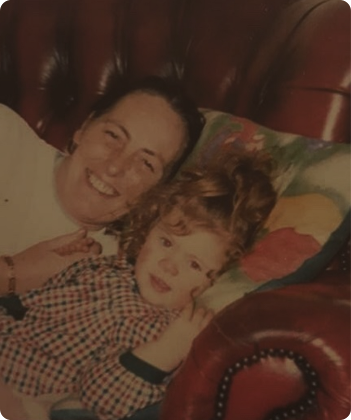 A woman and a young girl lying on a sofa, smiling and looking at the camera. The woman has short dark hair, and the girl has curly red hair. They are resting on a colourful pillow near a dark brown headboard.
