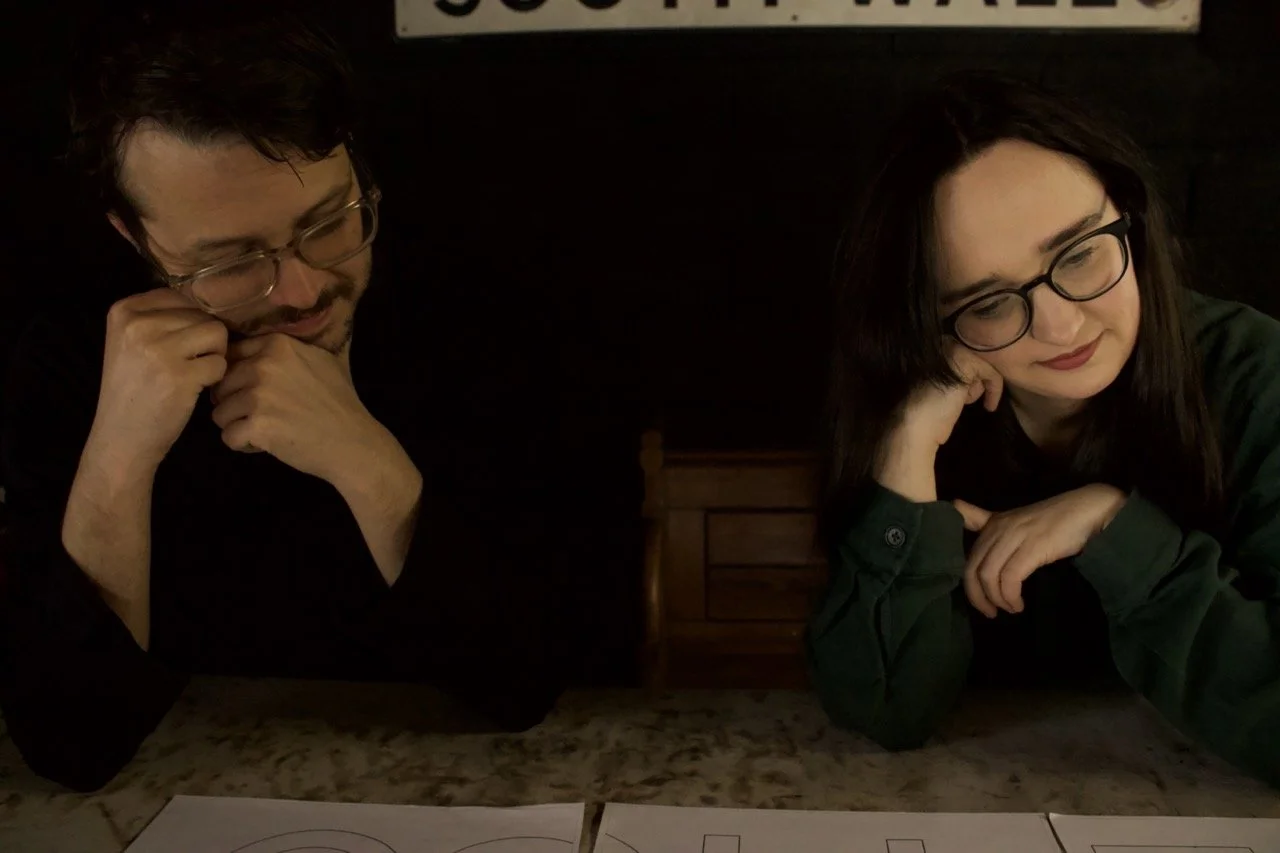 Two people sitting at a table, smiling and making playful faces. Behind them is a sign that reads 'South Wales.'
