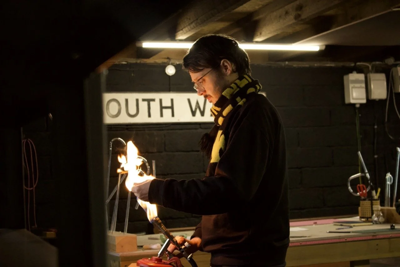 Neon maker bending a glass tube over an open flame inside a dark studio, wearing glasses and a striped scarf, with tools and a workbench visible in the background and a partially visible “South Wales” sign on the wall.