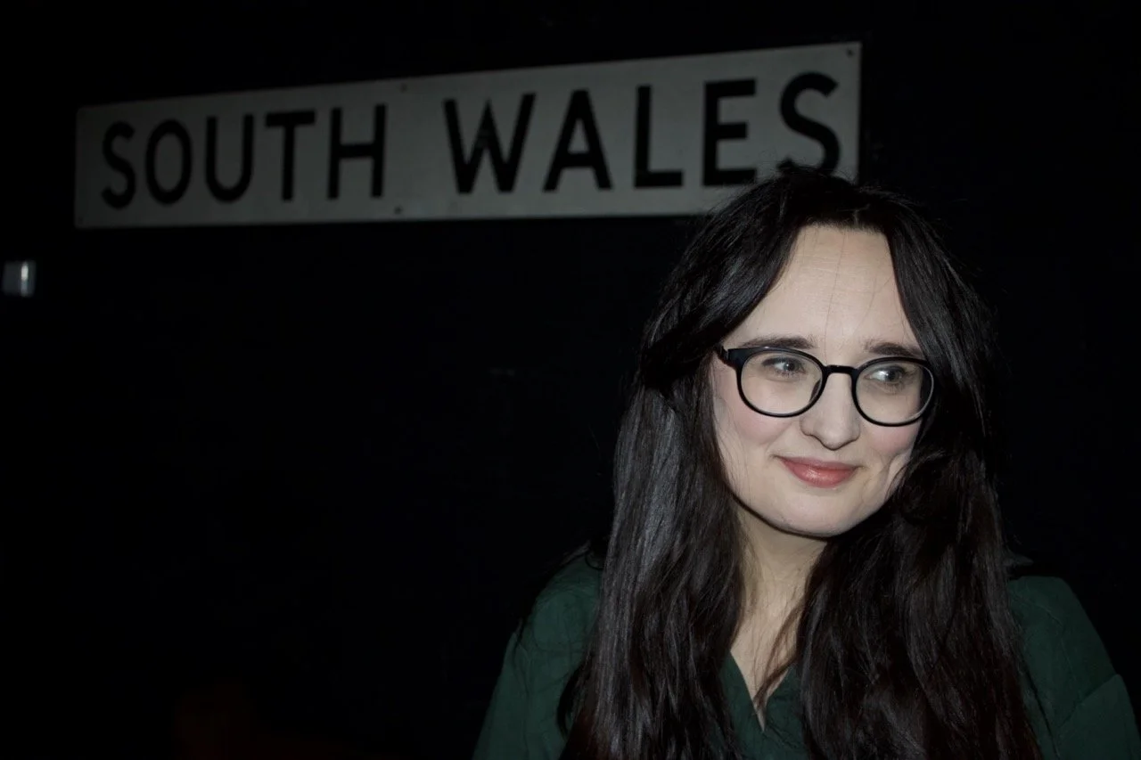 Portrait of a woman with long dark hair and round black glasses, softly lit against a dark background, standing in front of a vintage-style sign reading “South Wales.”