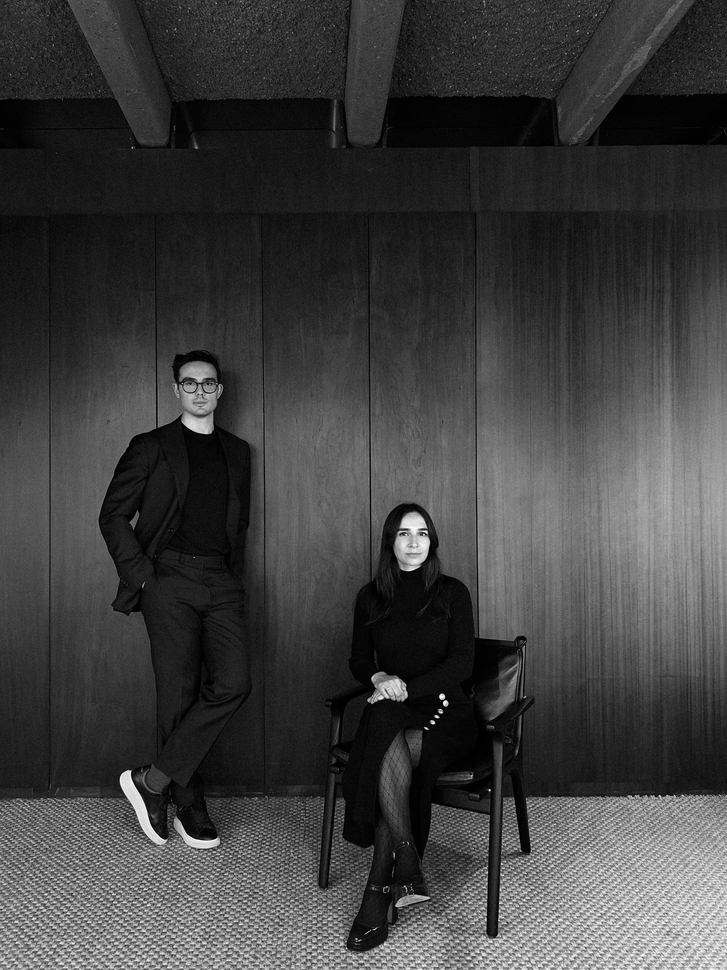 Deux personnes posant dans une pièce moderne, l'une debout et l'autre assise sur une chaise, toutes deux en vêtements sombres. Mur en bois en arrière-plan.