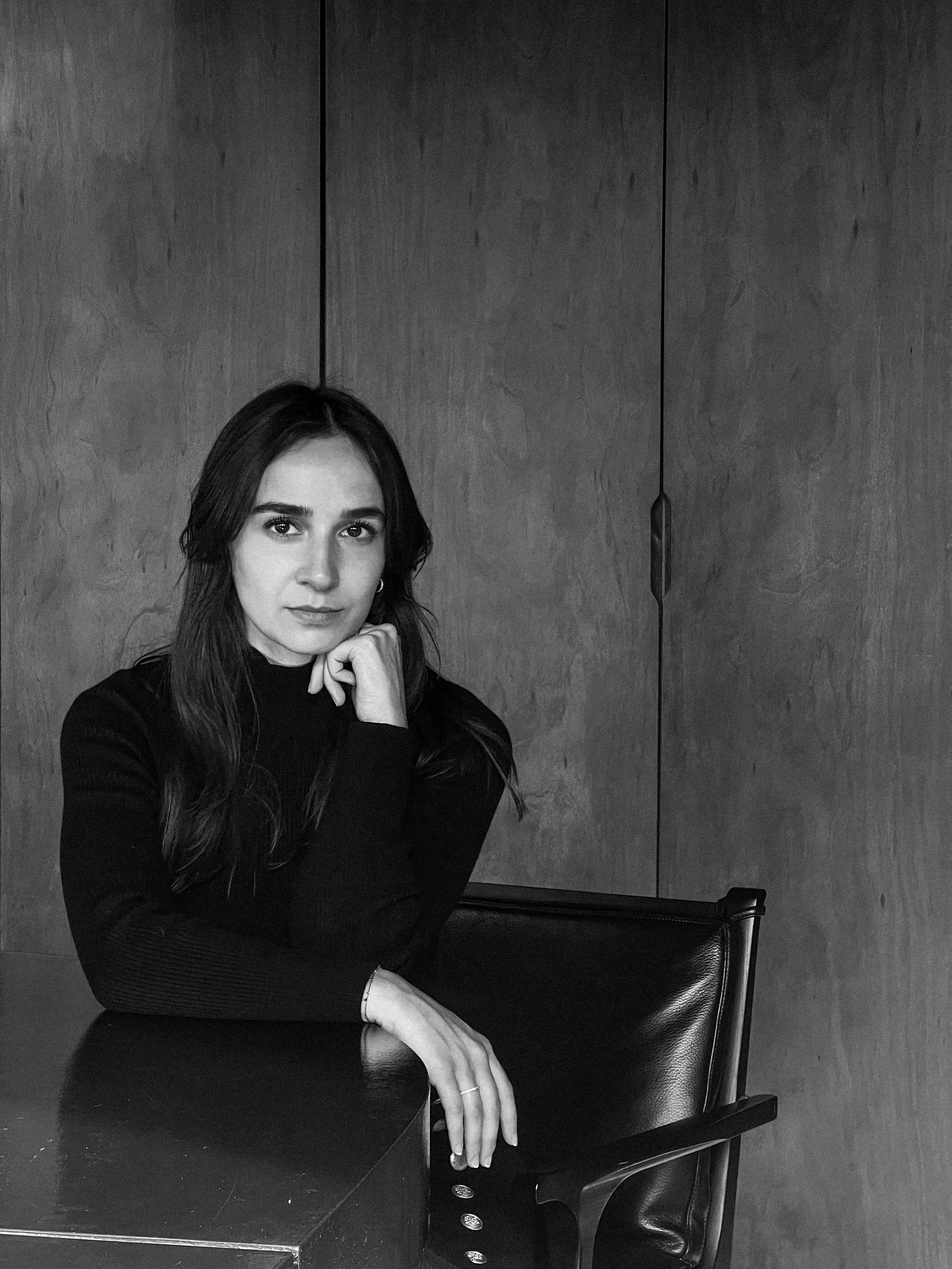 Femme assise à une table, portant un pull noir, avec un doigt posé sur le menton, en noir et blanc.