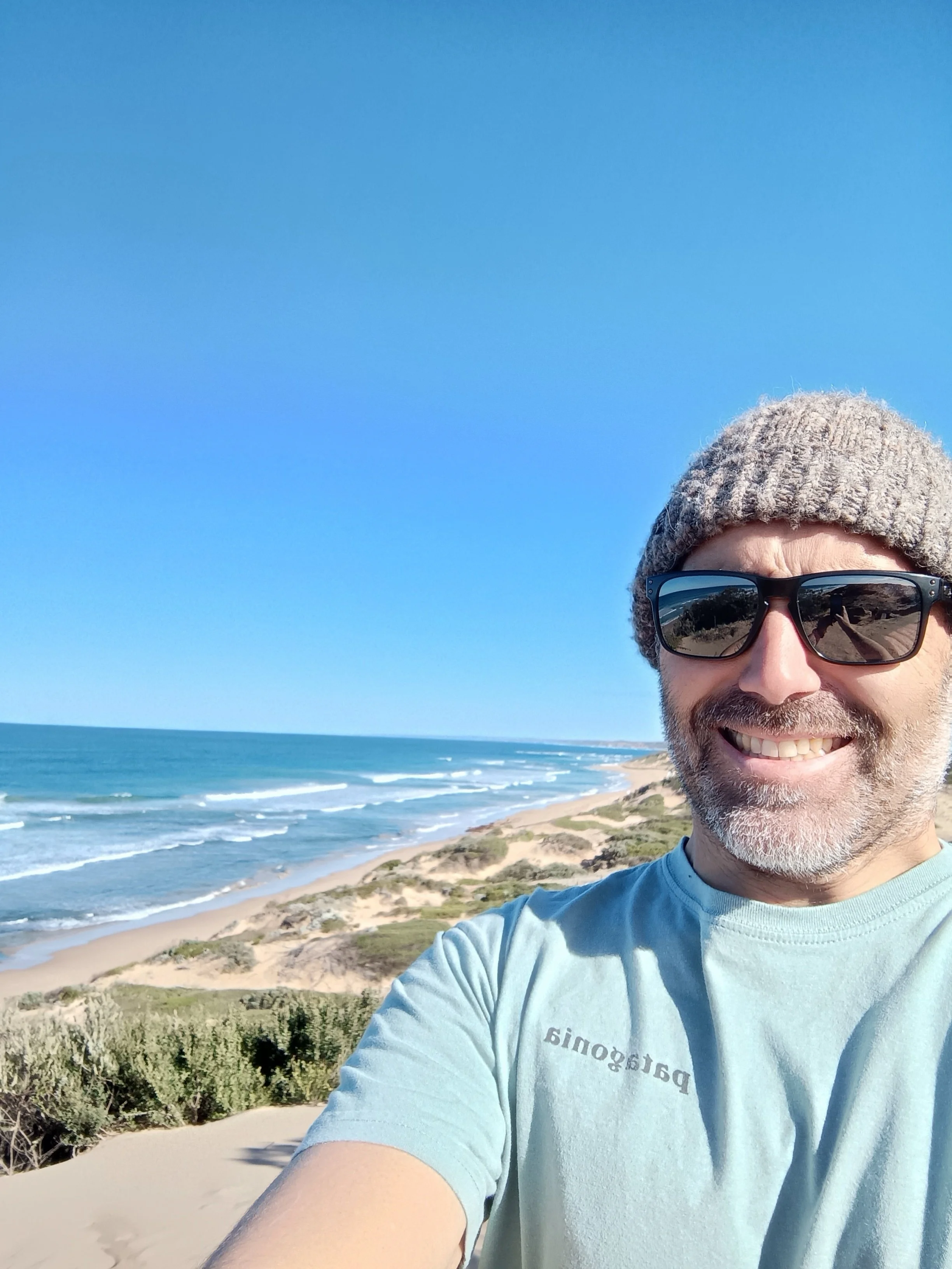 A smiling man taking a selfie at the beach, wearing sunglasses, a knit beanie, and a light blue t-shirt with partially visible text. The background features a sandy beach, ocean waves, greenery, and a clear blue sky.