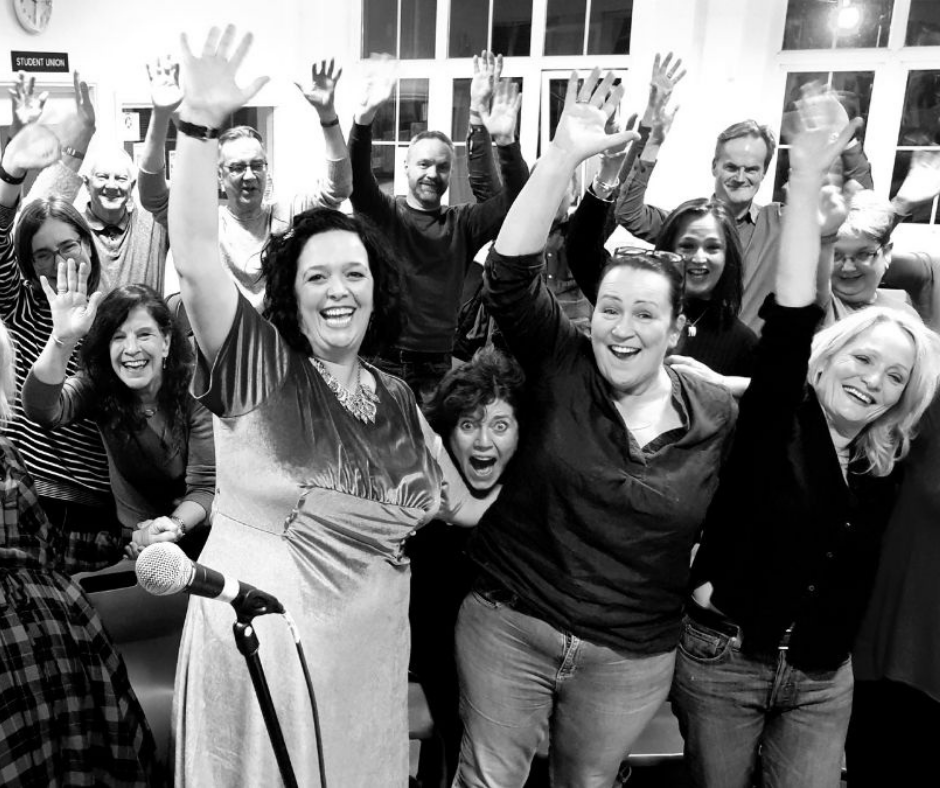 Bright Soul Choir members smiling and raising their hands in a room with large windows, some women are in the front, one woman is speaking into a microphone, all appearing joyful.