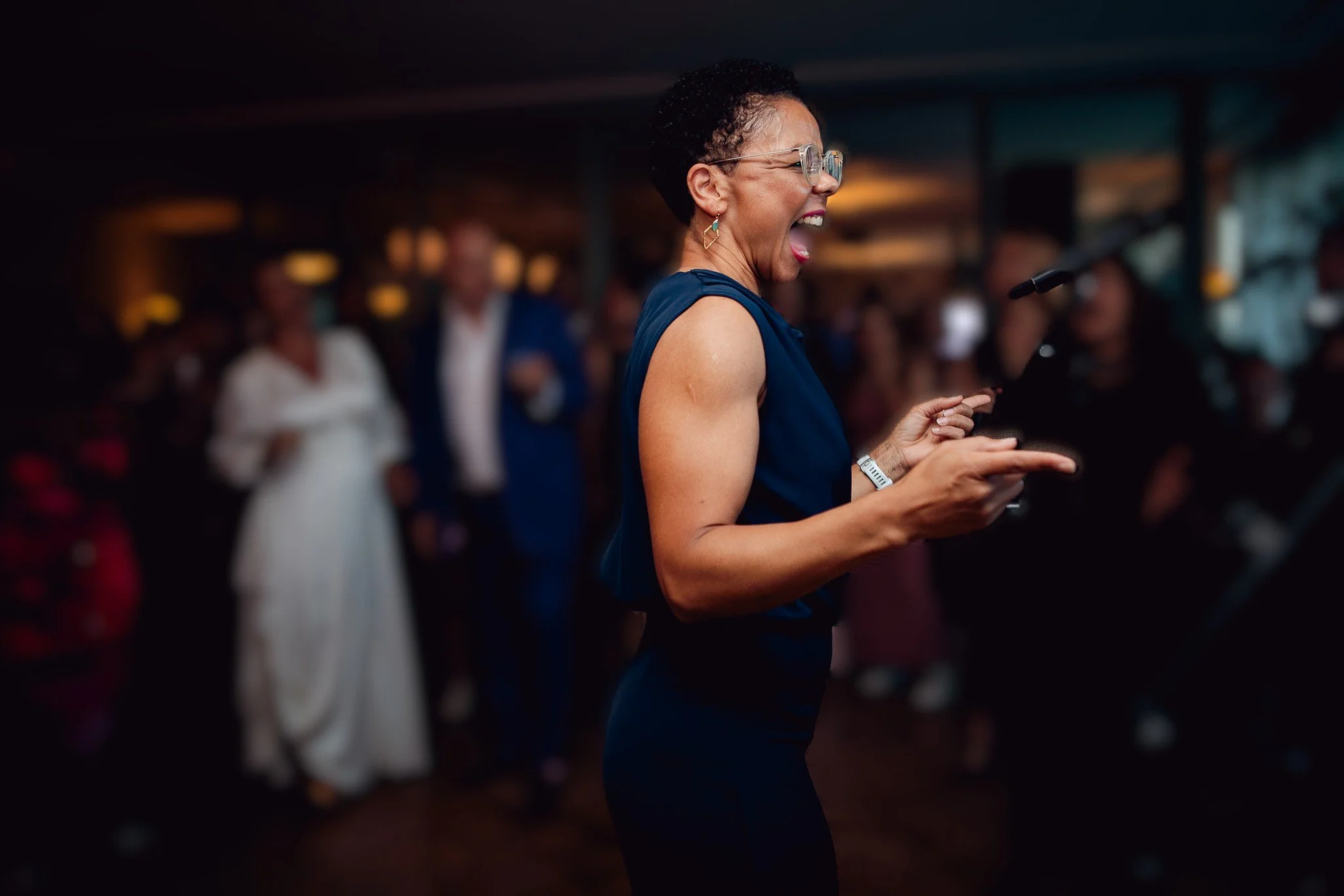 Choir leader, Amanda, in a dark blue dress is dancing and smiling at a wedding event, with blurred guests in the background.