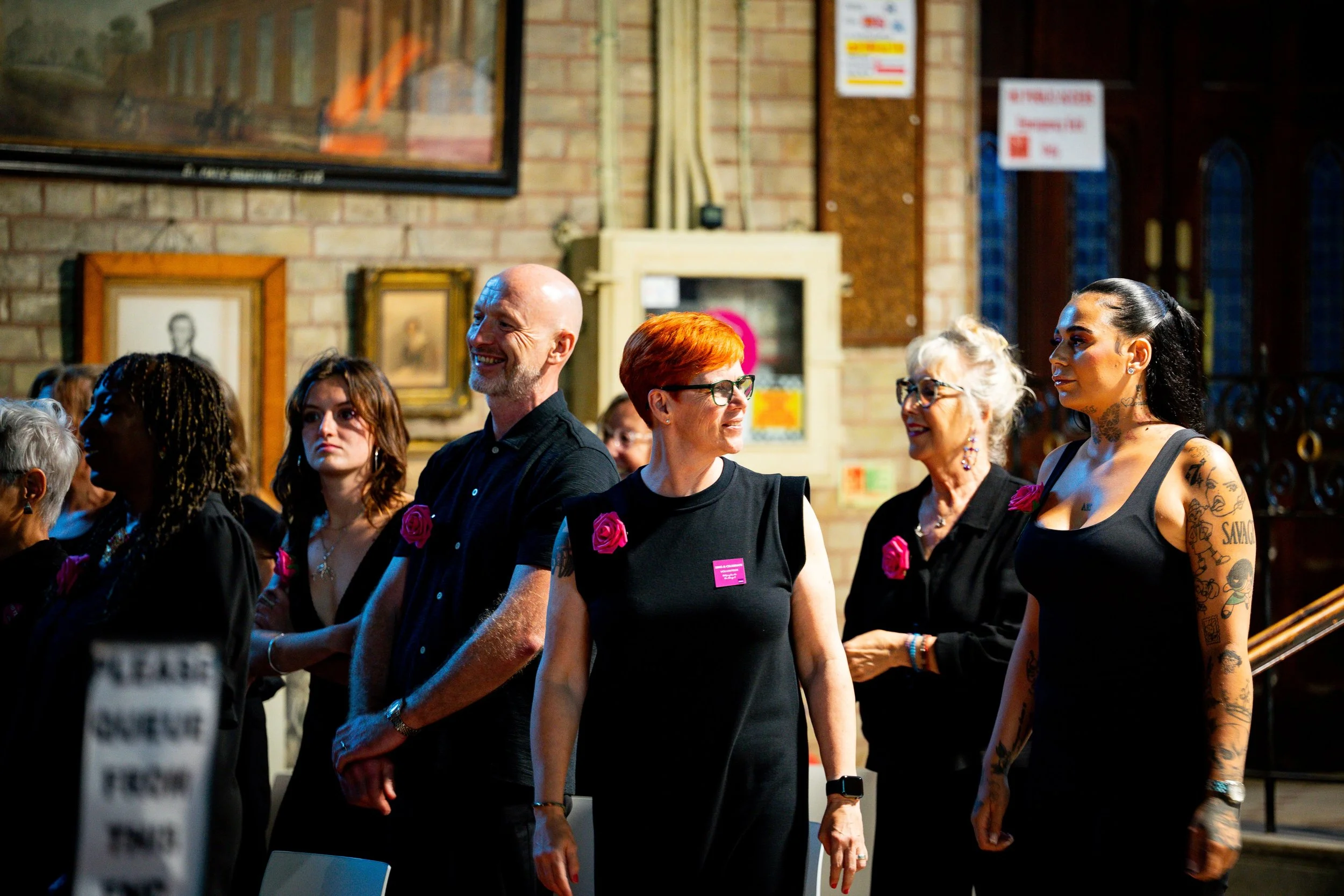 Choir members smiling together at a concert. Each one wearing a black outfit with a magenta rose