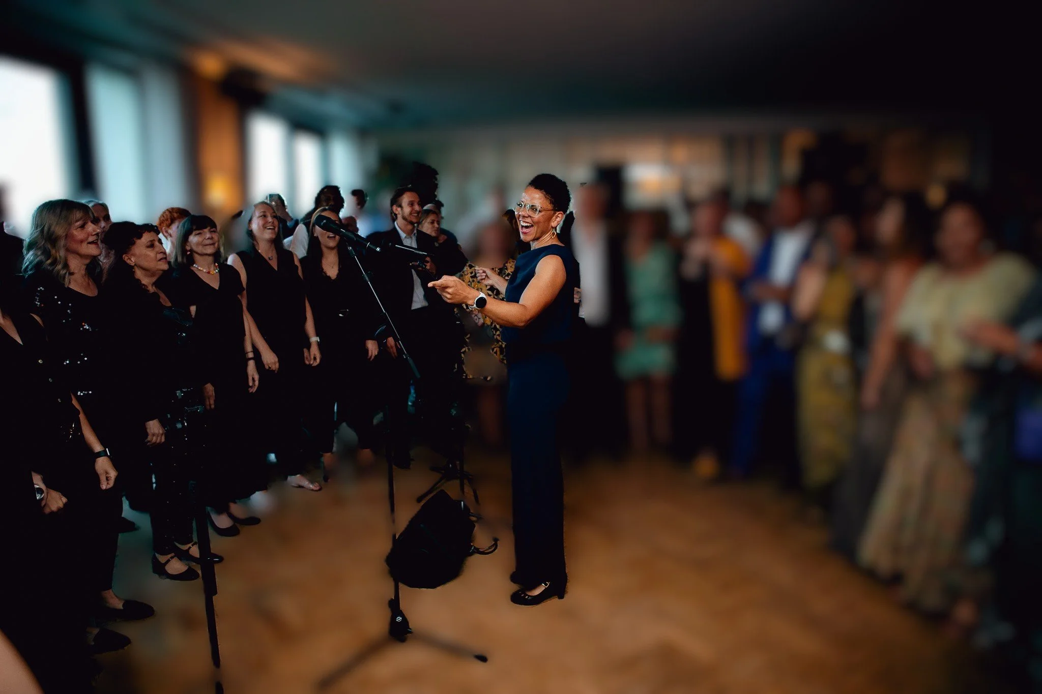 Choir Leader Amanda, in blue dress singing with choir and audience in a room with large windows.