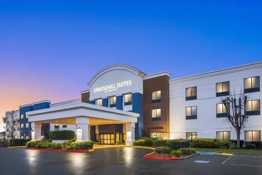 Exterior view of SpringHill Suites hotel featuring a modern three-story building with a covered entrance, landscaped grounds, and illuminated façade at dusk.