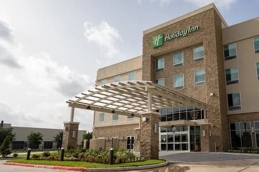 Exterior view of a Holiday Inn hotel featuring a modern multi-story building with a stone façade, covered entrance canopy, landscaped grounds, and main glass entry doors.