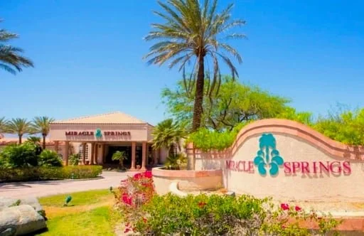 Entrance to Miracle Springs Resort featuring a desert-style landscape with palm trees, flowering plants, and a monument sign in front of the main building under a clear blue sky.