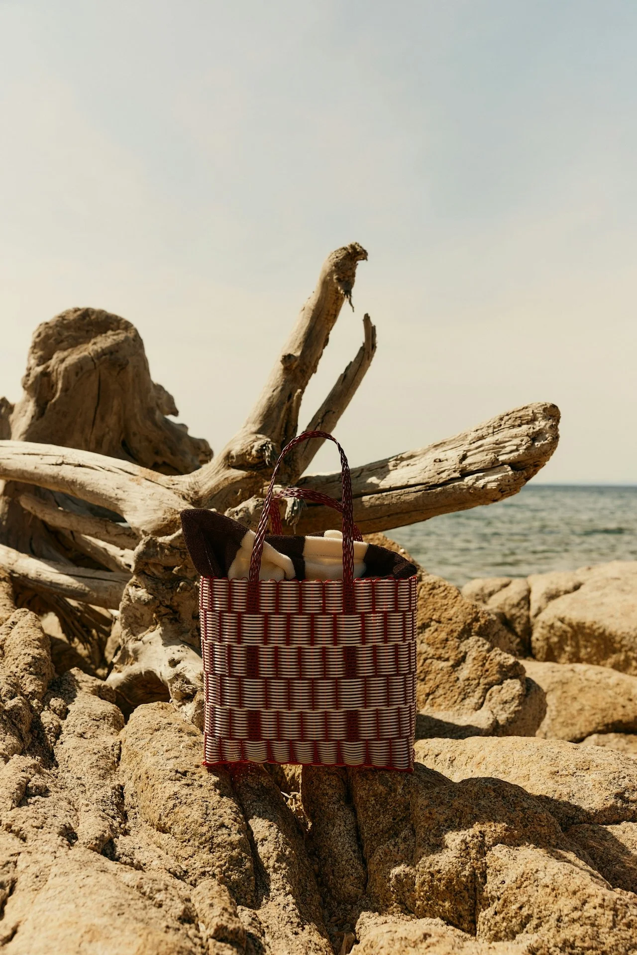 A woven red and white basket with items inside, placed on rocks near a driftwood branch by the sea, under a pale blue sky.