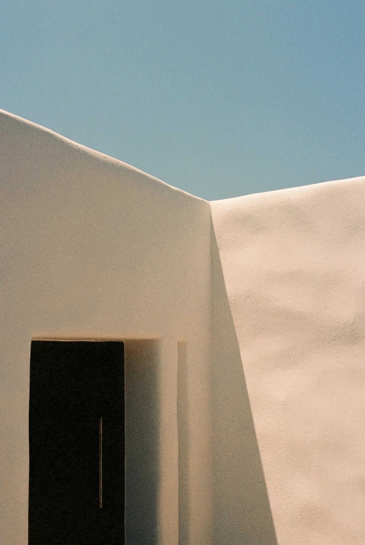 Minimalist white building with a curved roof and a dark rectangular opening, against a clear blue sky.