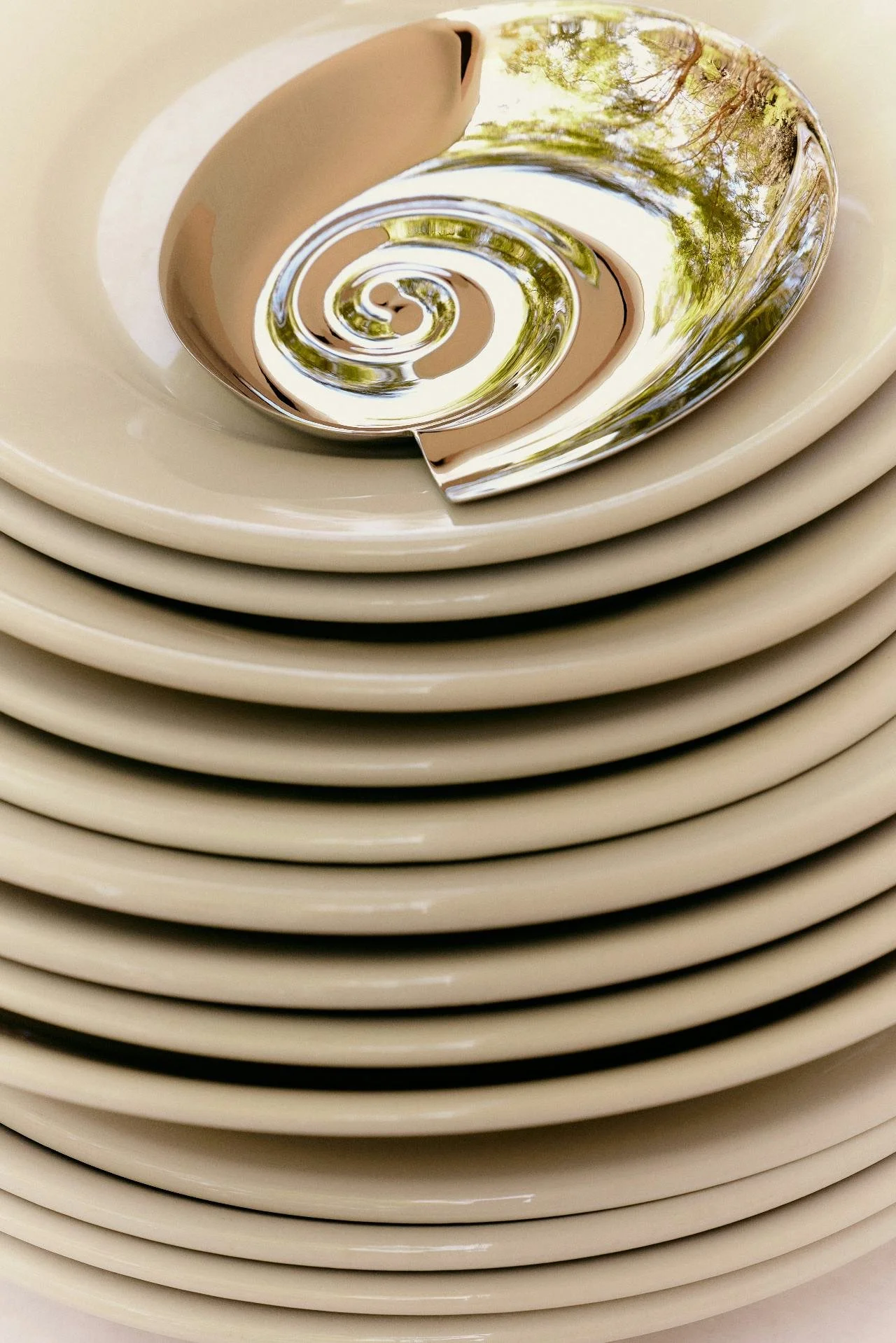 A metallic spiral sculpture reflecting trees and sky, placed on a beige stack of plates.