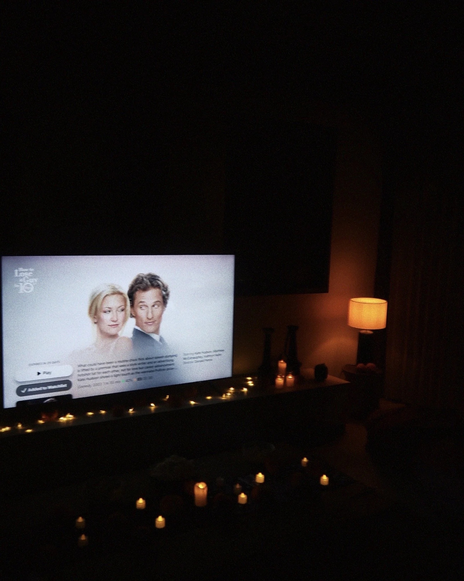 Living room with a television displaying a movie or show, a lamp emitting warm light, candles, and decorative vases on a table.
