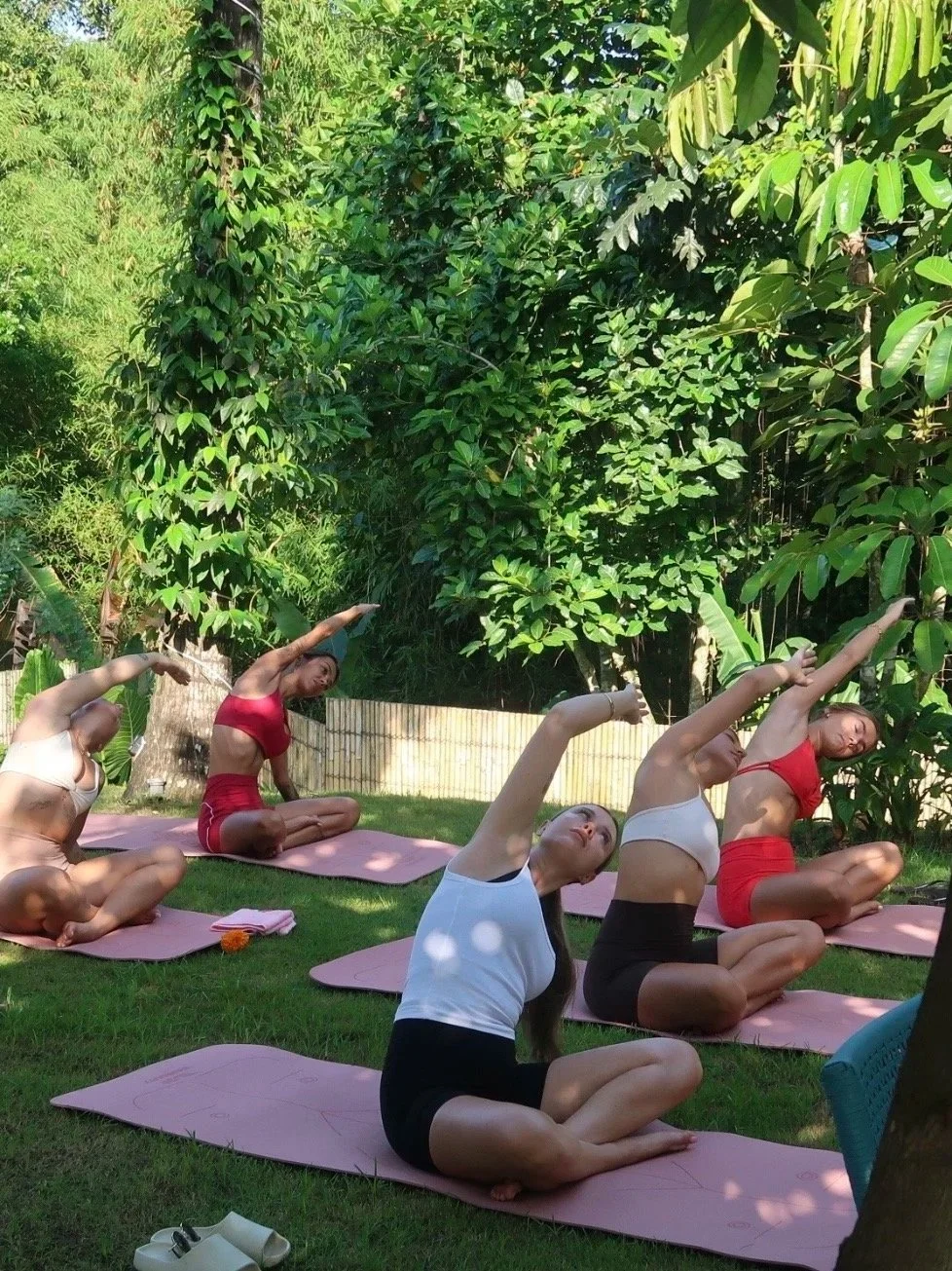 Group of people practicing yoga outdoors on pink mats in a garden with lush green trees and plants.