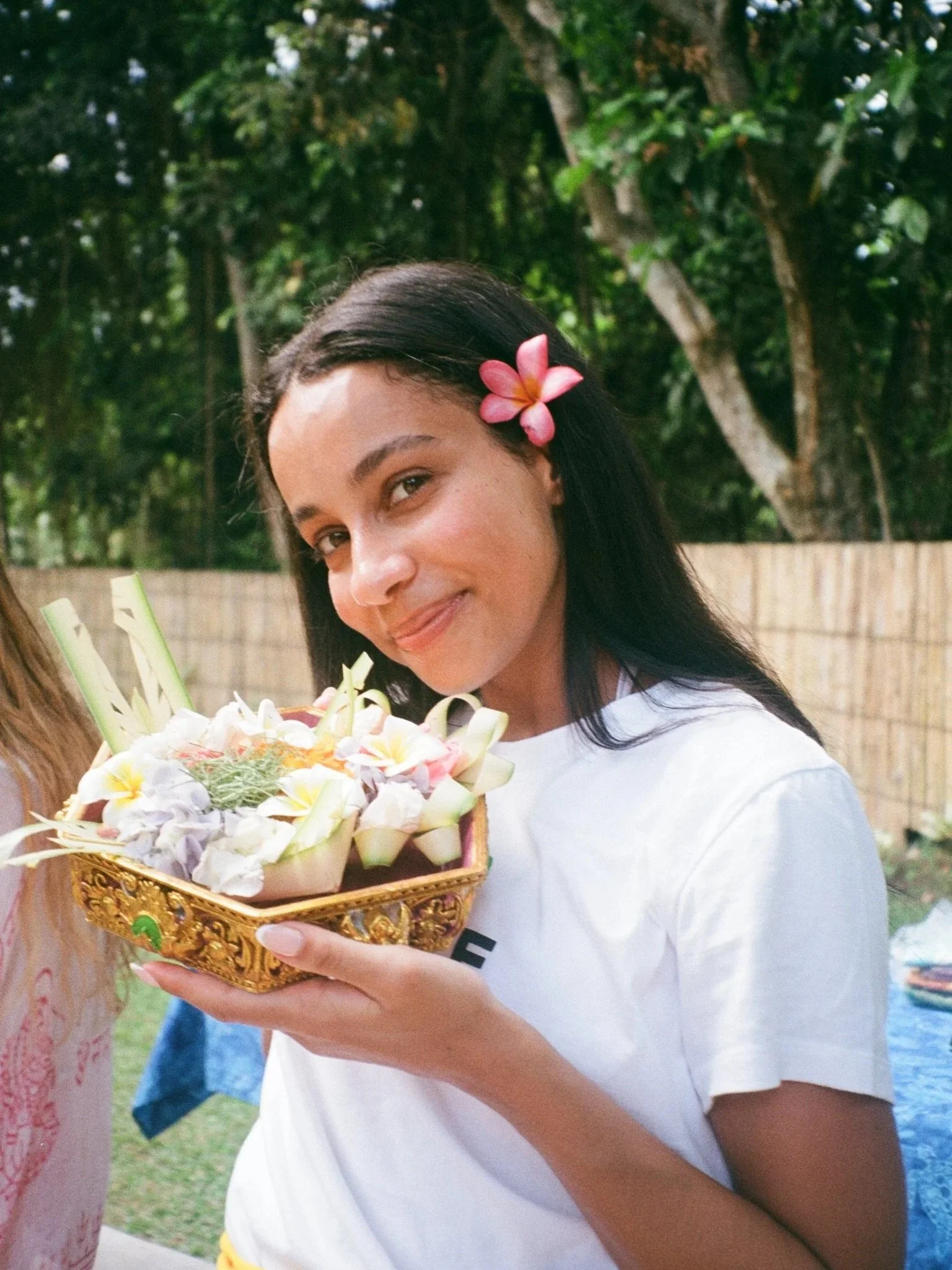 Young woman with dark hair, wearing a white shirt and a pink flower in her hair, holding a decorative bowl filled with flowers and greenery at an outdoor setting with trees and a bamboo fence in the background.