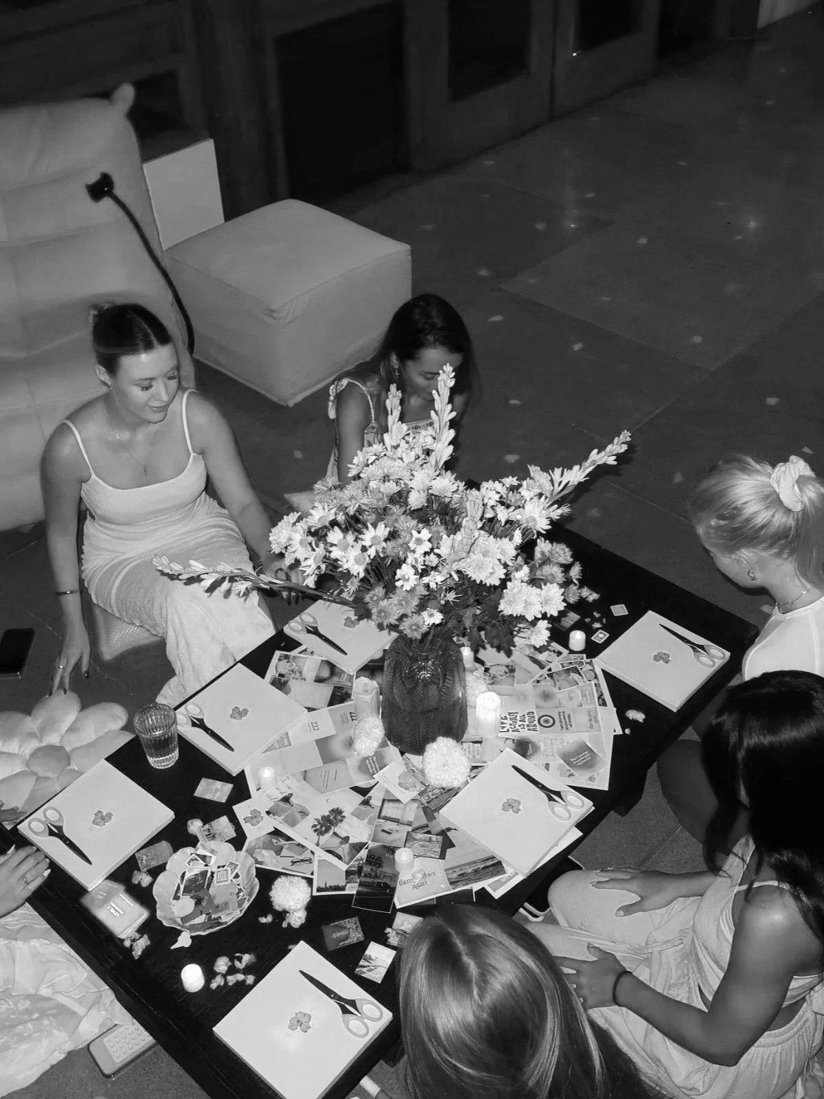 A black-and-white photo of five women gathered around a table decorated with a large flower centerpiece and scattered papers, scissors, and candles, possibly engaged in a crafting or flower arranging activity.
