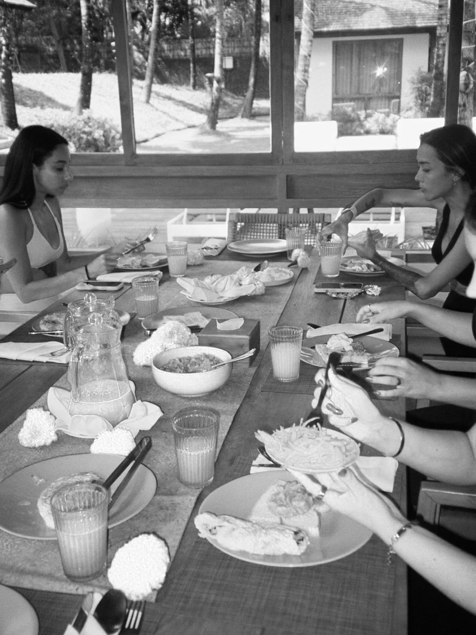 Women sitting around a dining table with food, drinks, and utensils, inside a room with windows showing trees outside.