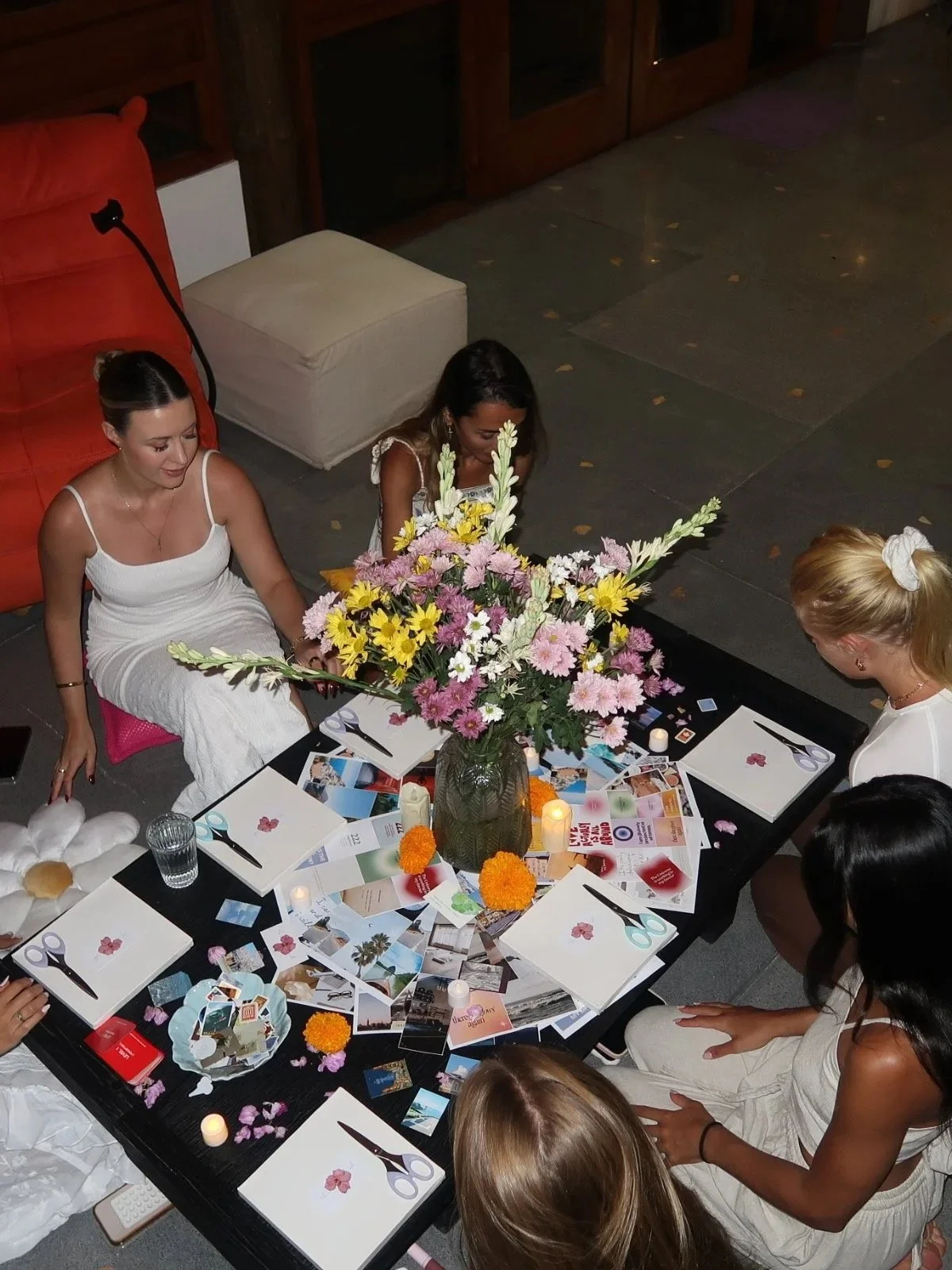 A group of women sitting around a table decorated with a large flower bouquet, candles, and assorted photos and papers, engaged in a social or celebratory activity.