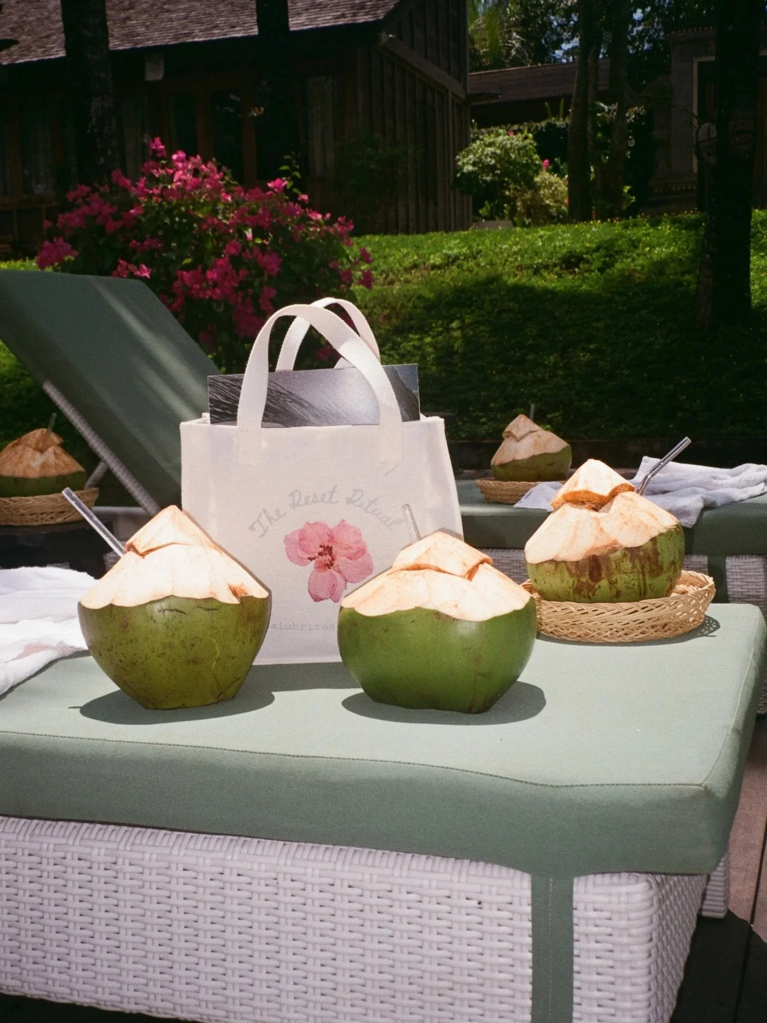 Three young coconuts with their tops shaved off, placed on a green cushioned outdoor table, with some spoons inserted. In the background, there is a white bag with a pink flower design, a lounge chair, and a lush garden with flowers and trees.