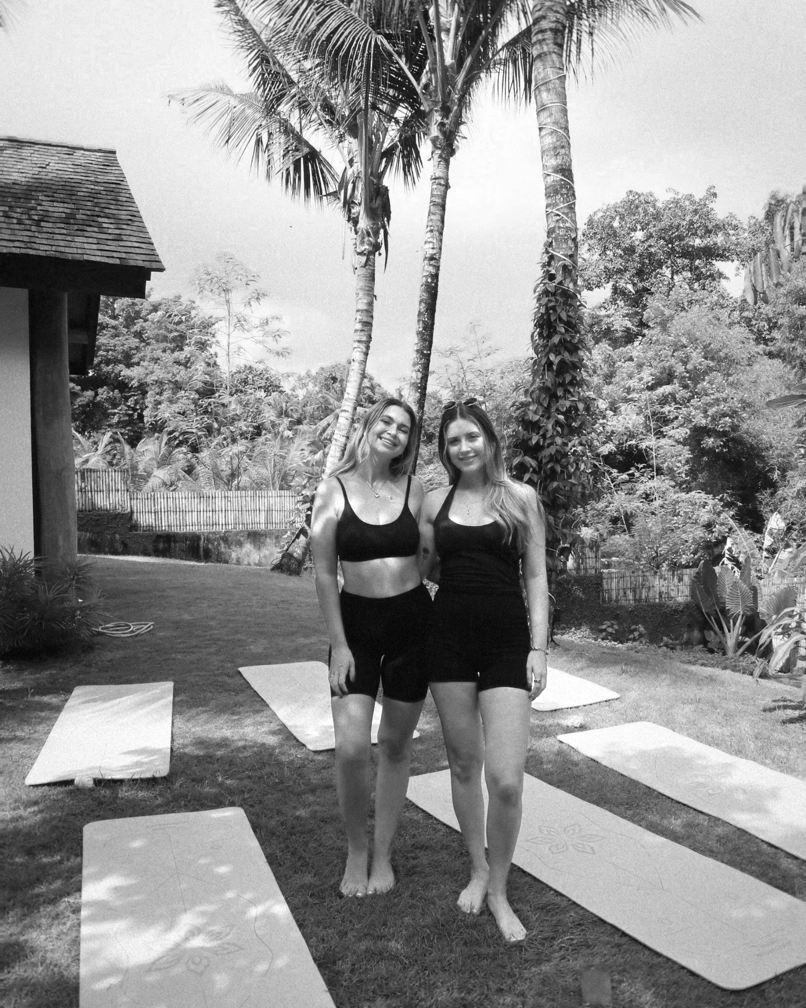 Two women in black workout attire standing on yoga mats in an outdoor garden with tall palm trees and lush foliage in the background, smiling at the camera.
