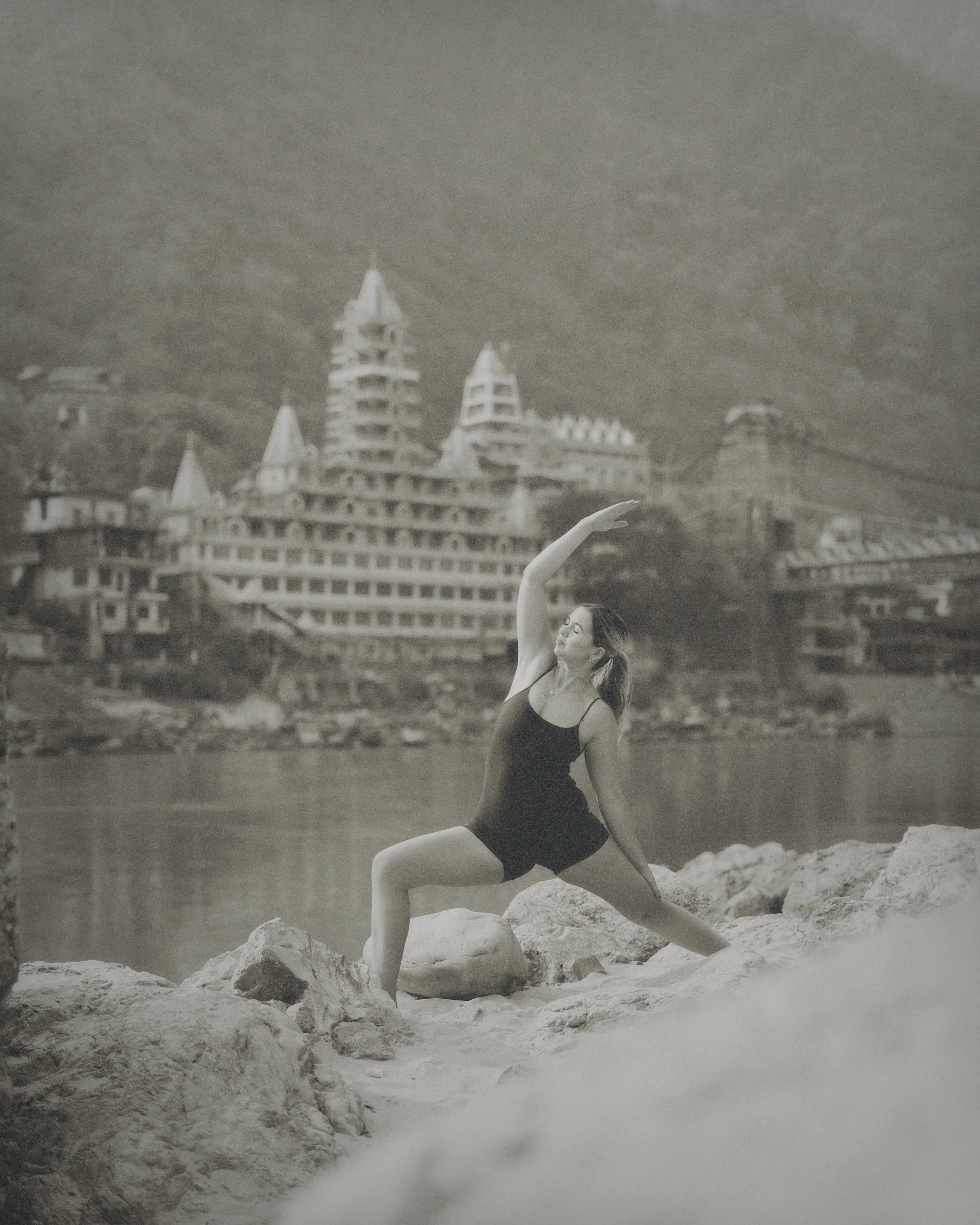 A woman practicing yoga on rocks by the water with a large building and mountains in the background.
