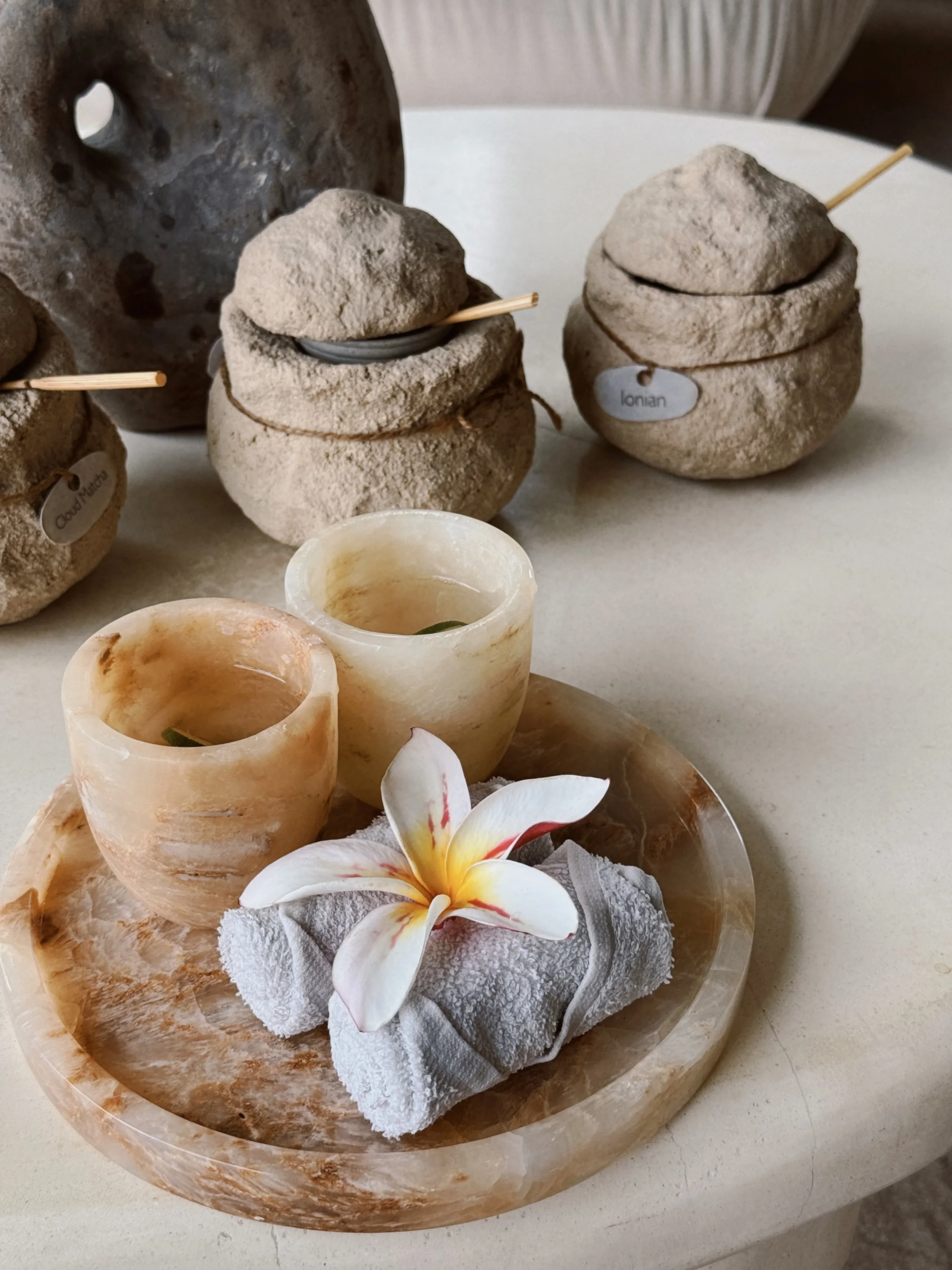 Beach-themed spa arrangement with two small orange onyx candle holders, white towel, and a flower on a round marble tray, with sandy rocks and labeled jars in the background.