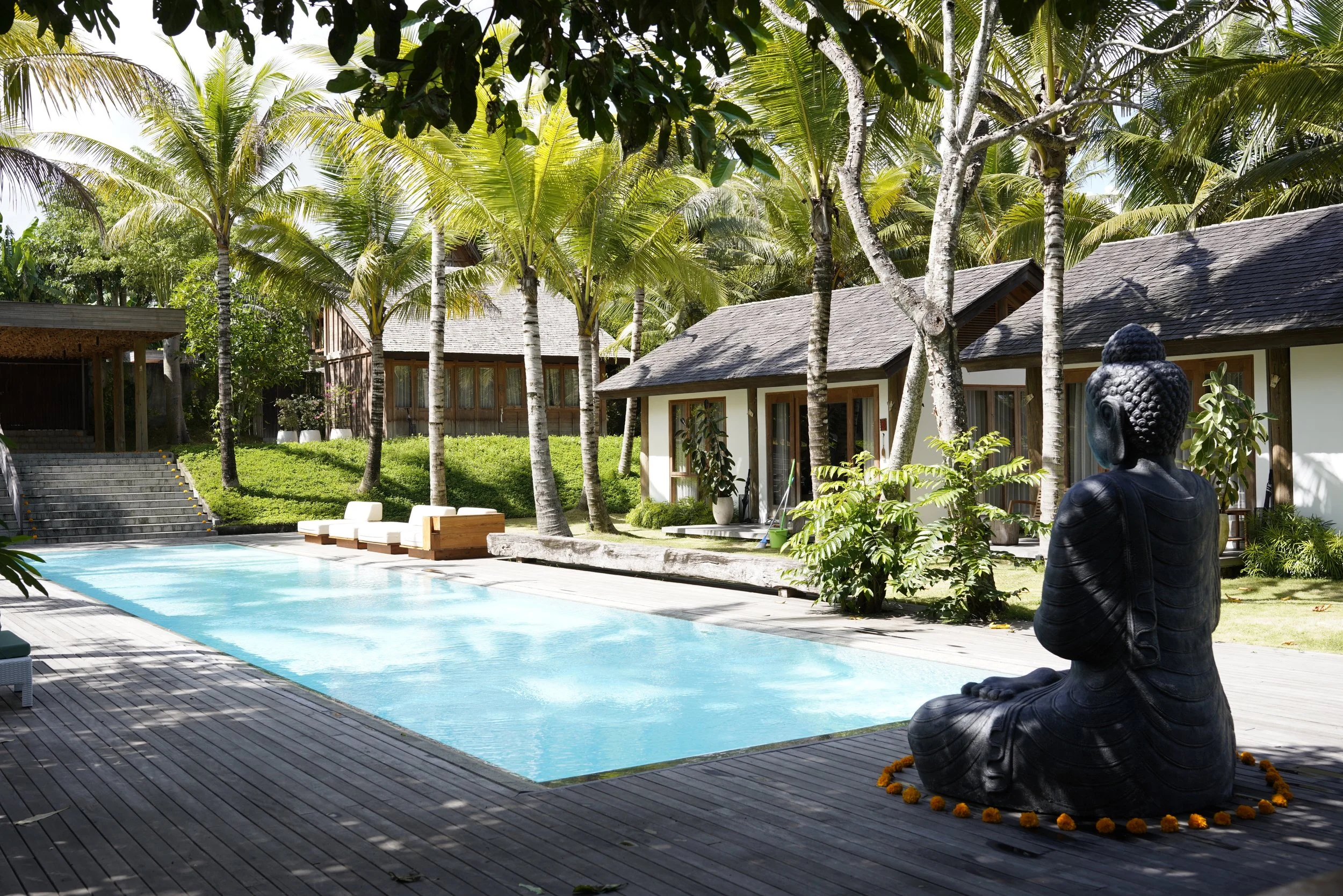 A serene outdoor scene featuring a rectangular swimming pool, surrounded by wooden decking, tropical trees, and small cottages. A seated black Buddha statue decorated with orange marigold flowers is in the foreground.