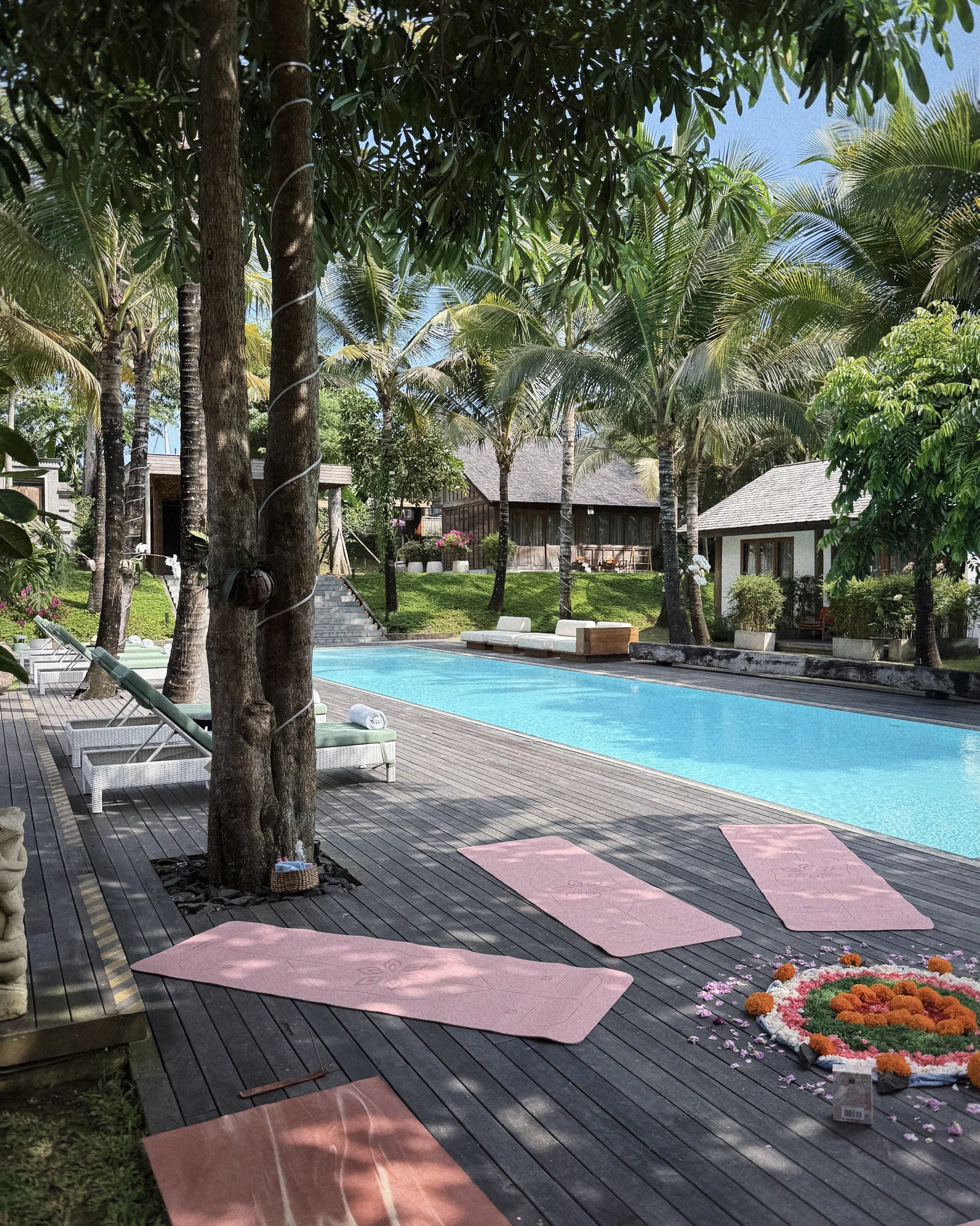 A serene outdoor pool area surrounded by palm trees and lounge chairs with pink yoga mats set up on a wooden deck. There are flower arrangements on the deck and nearby buildings.