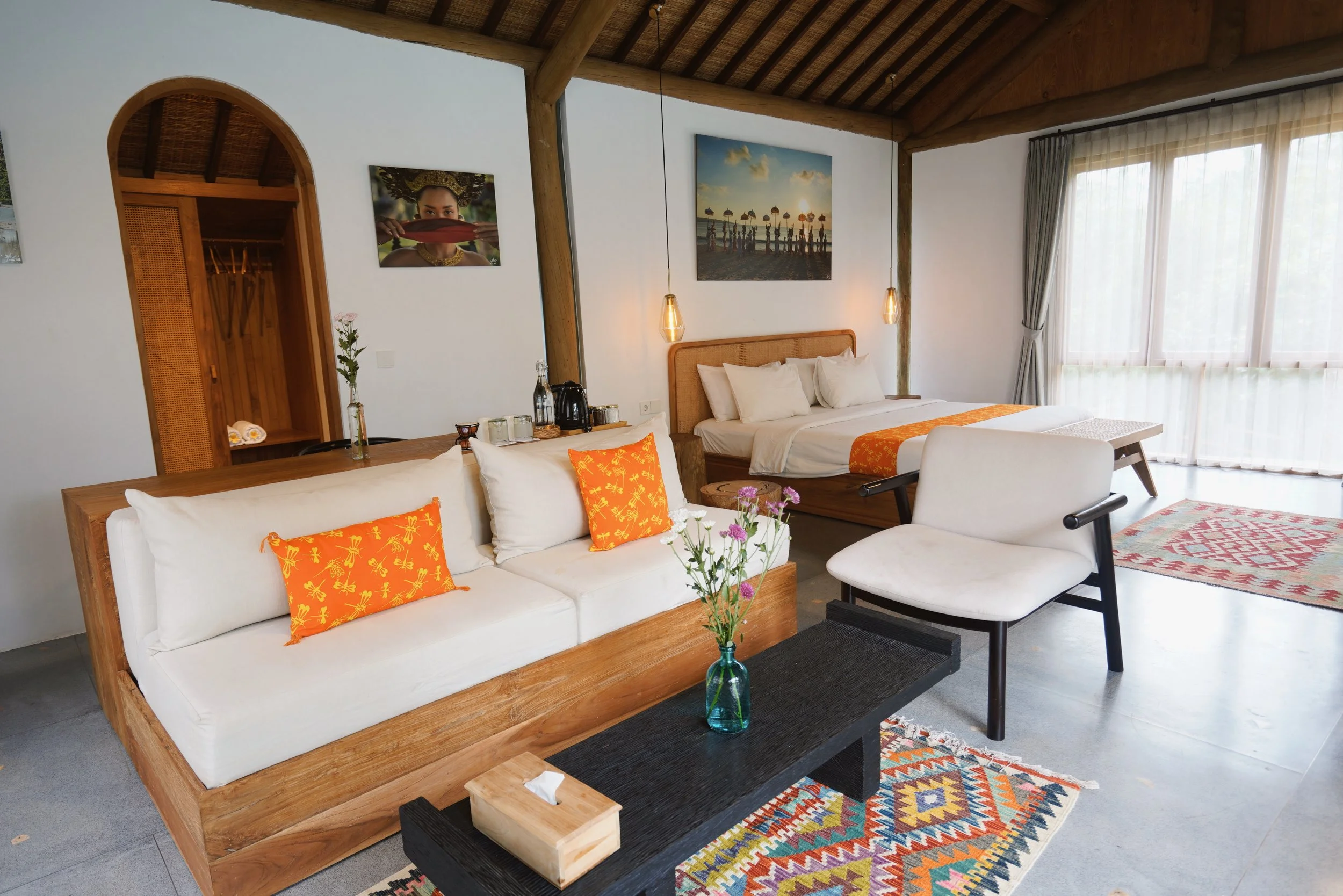 A spacious hotel room with a wood-beamed ceiling, a white sofa with orange patterned pillows, a wooden bed with white bedding and orange accents, a black table with a vase of purple flowers, a white armchair, and large windows with sheer curtains all