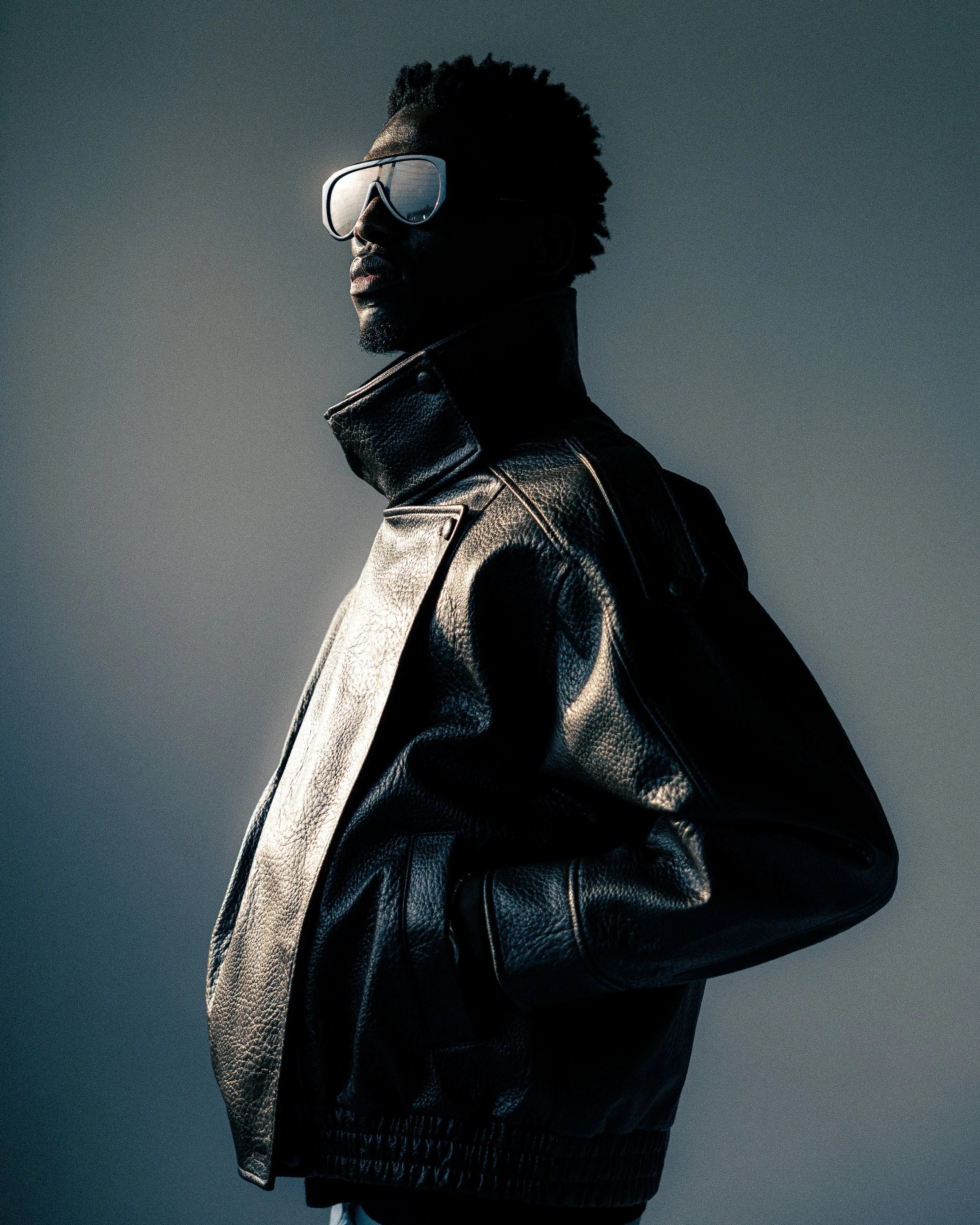 A person with curly hair wearing large goggles, a black leather jacket, and pants, standing against a dark, cloudy sky.