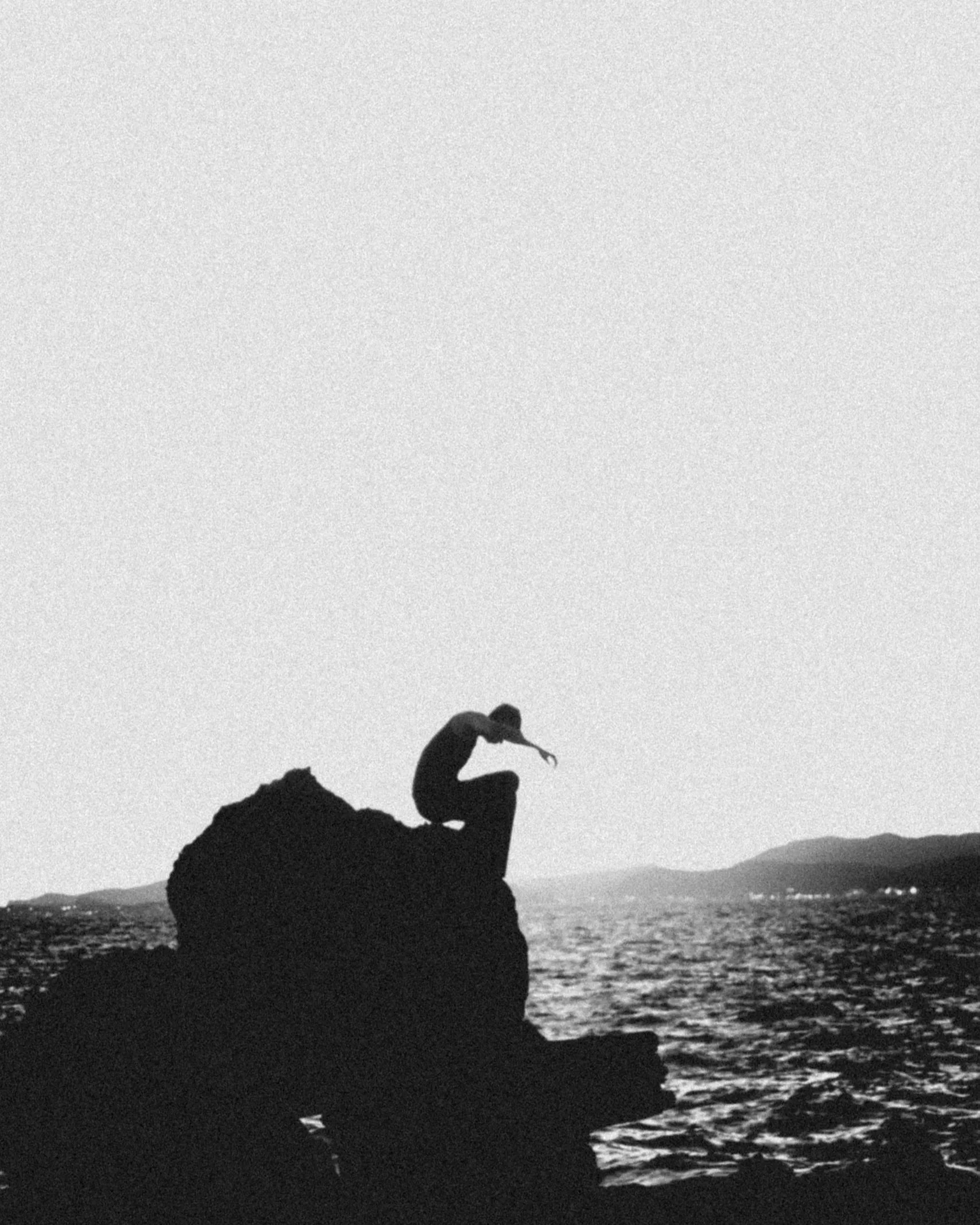 Silhouette of a person sitting on a rock by the water during sunset or sunrise with the sky and water reflecting light.