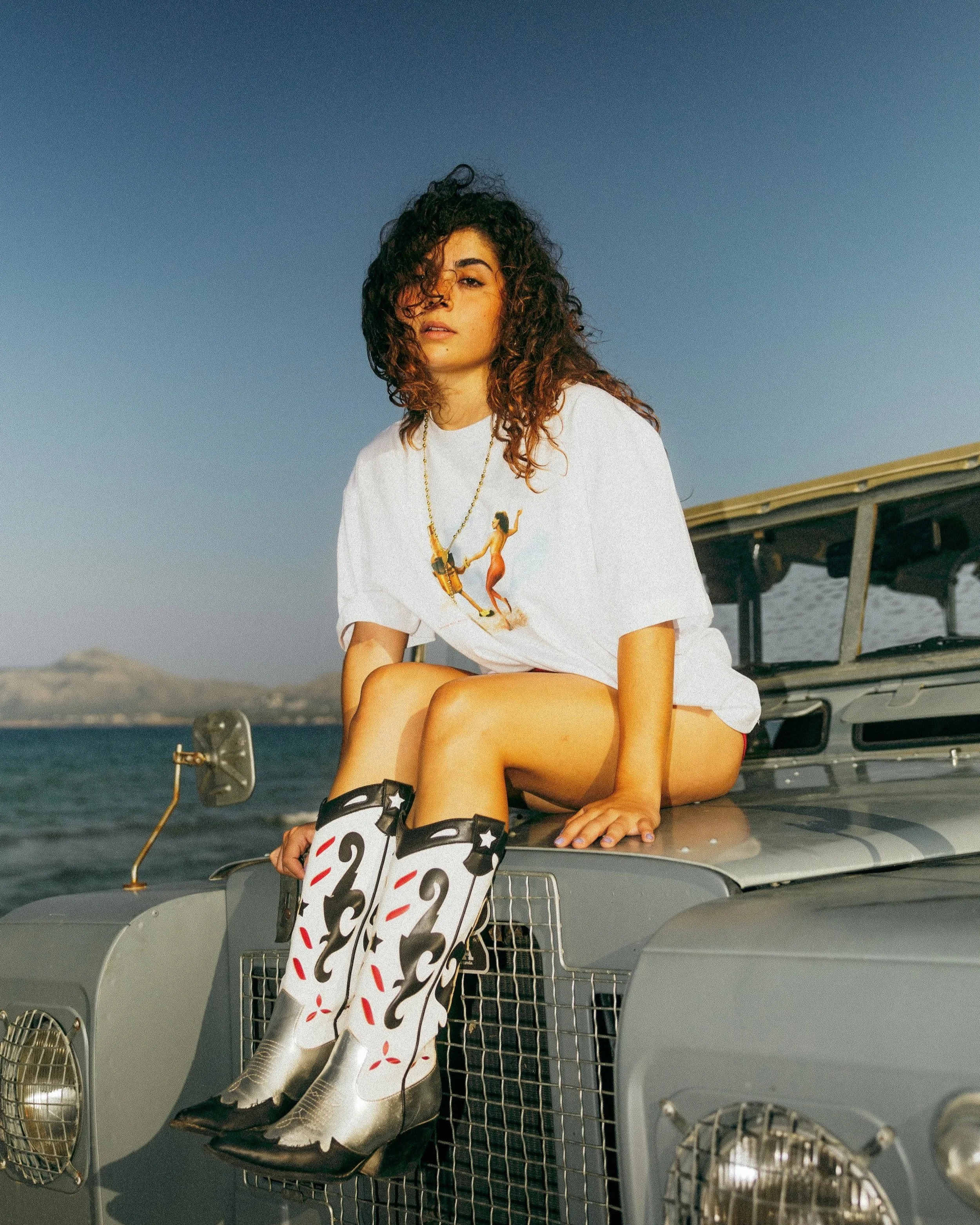 A woman with curly hair sitting on the hood of a military-style vehicle by the water, wearing a graphic t-shirt, cowboy boots with a black and white pattern, and a gold chain necklace.