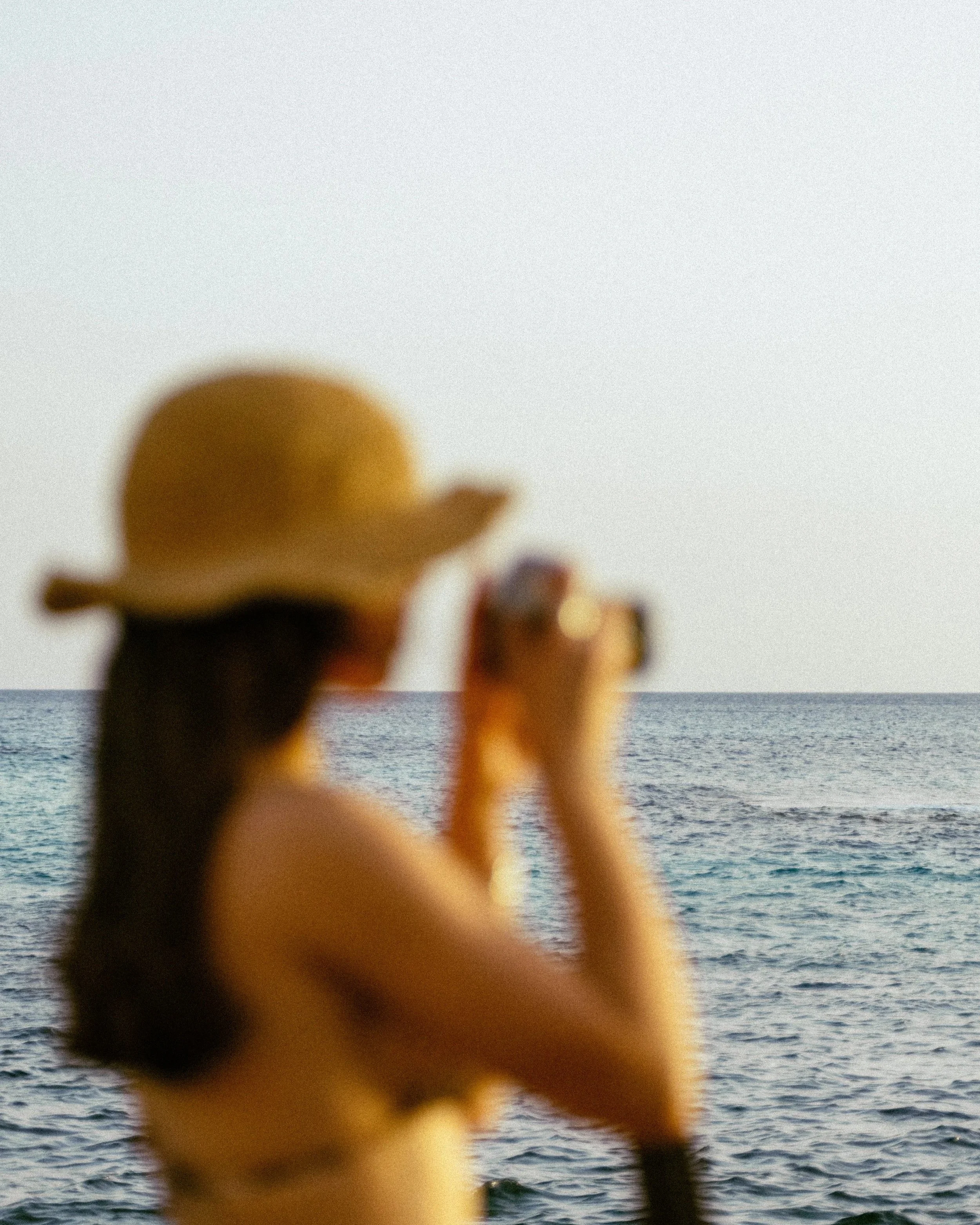 A woman wearing a sunhat standing by the ocean and taking a photo with her camera.