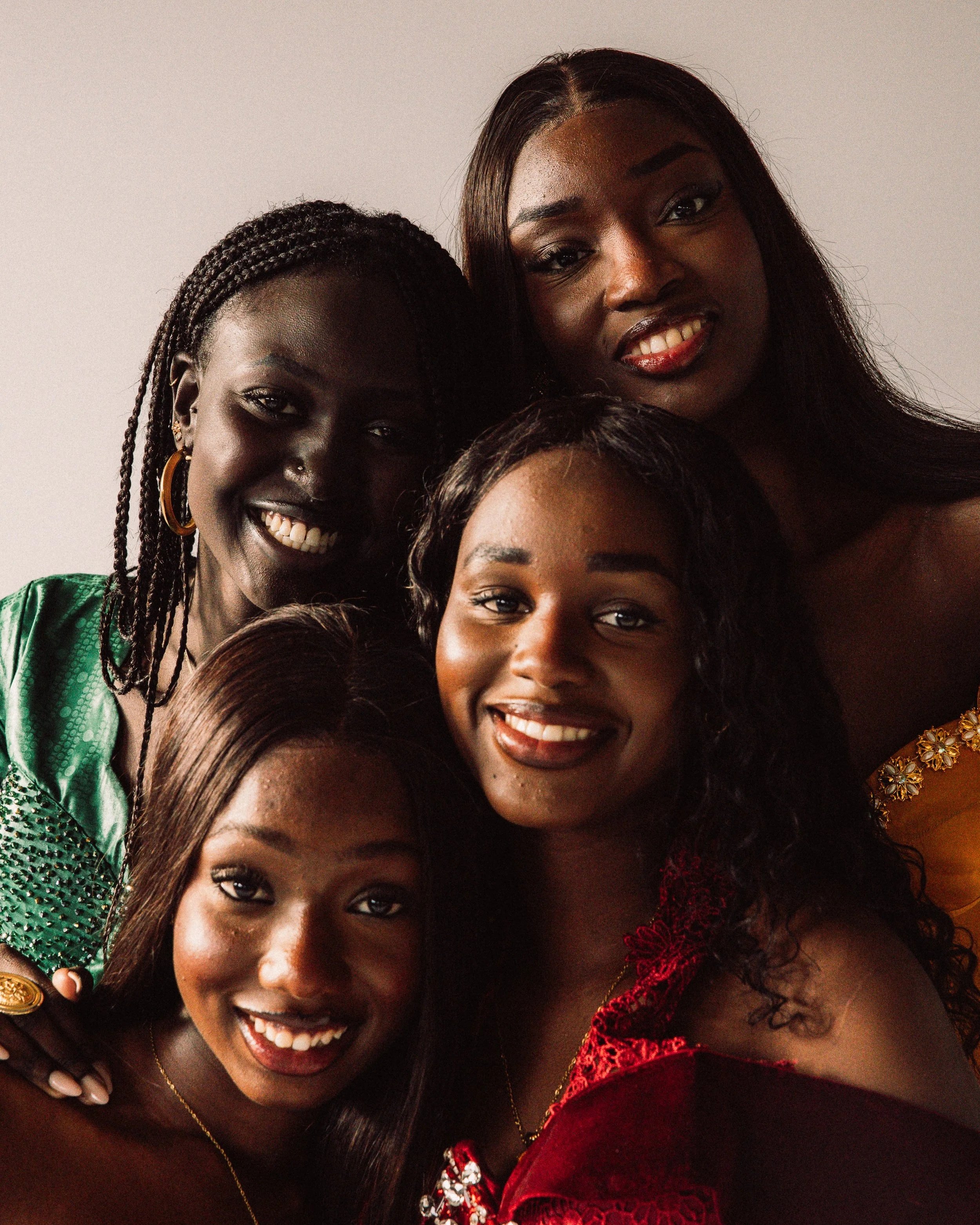 A group of women smiling and closely posing together, wearing colorful and elegant clothing, with a plain white background.