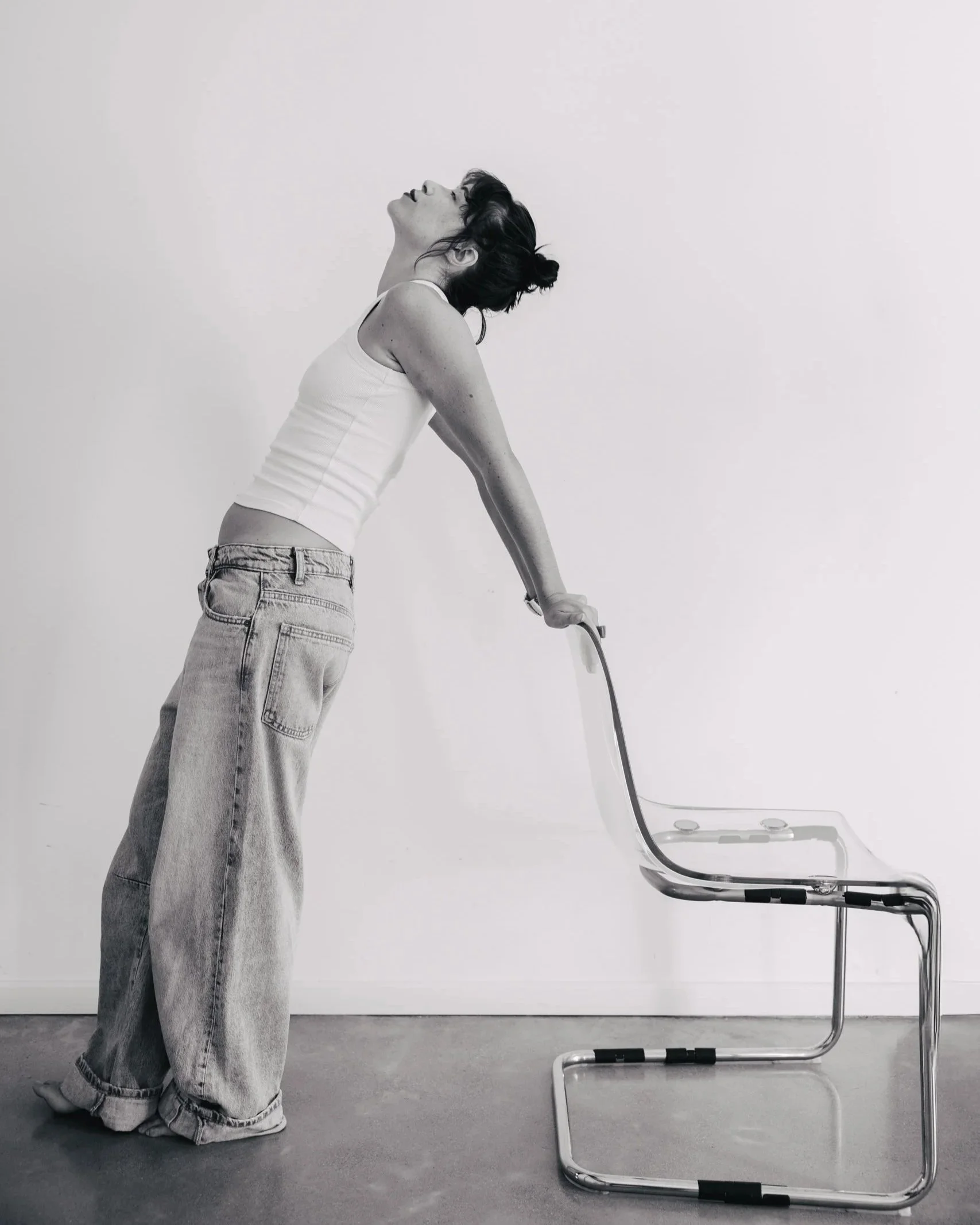 A person with dark hair, wearing a white tank top and loose jeans, is leaning back with their head tilted upward while holding a vacuum cleaner. The vacuum is placed on the ground, and the person appears relaxed or in a state of stretching.
