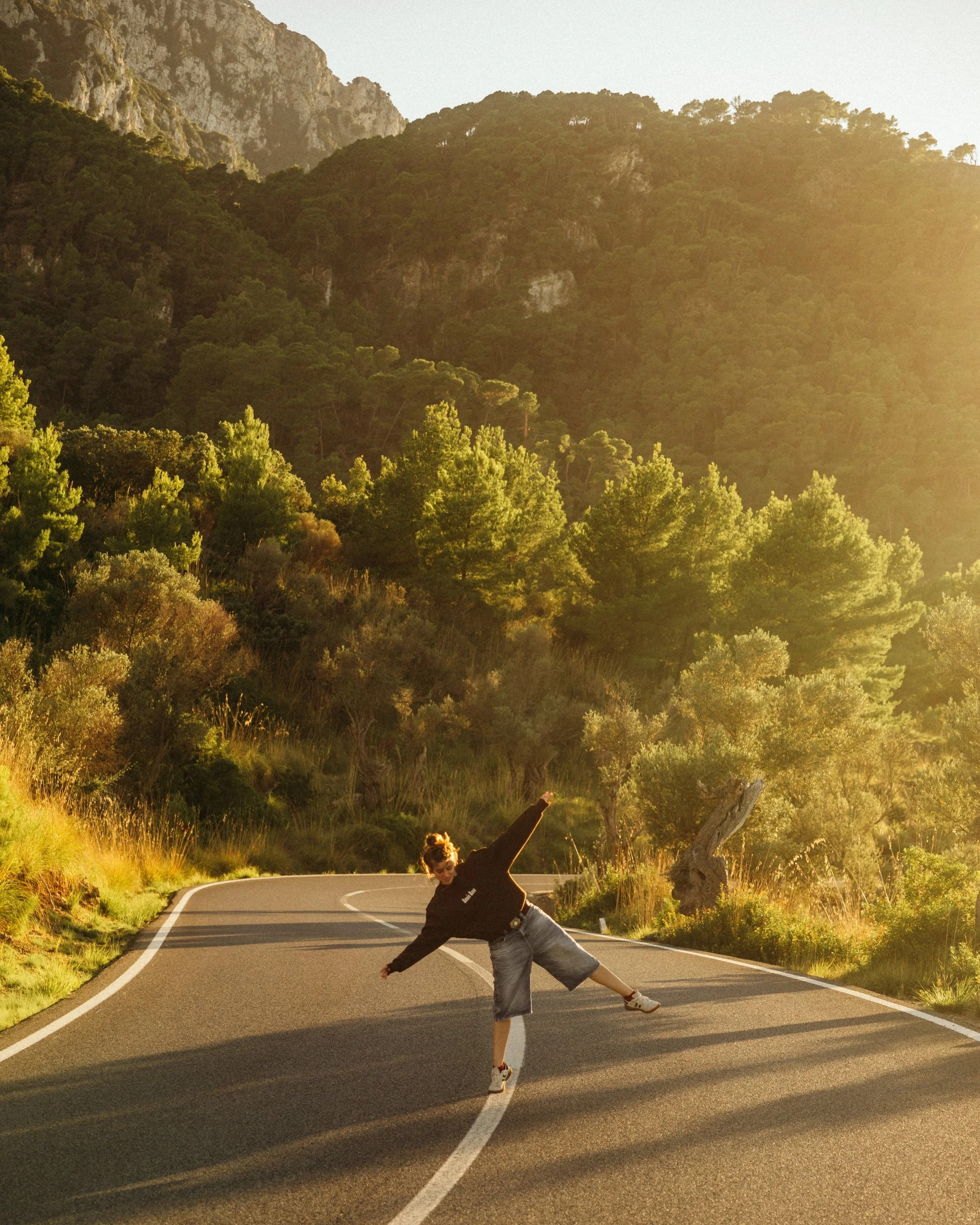 A person walking on a winding mountain road during sunset, surrounded by lush green trees and hills, with mountains in the background.