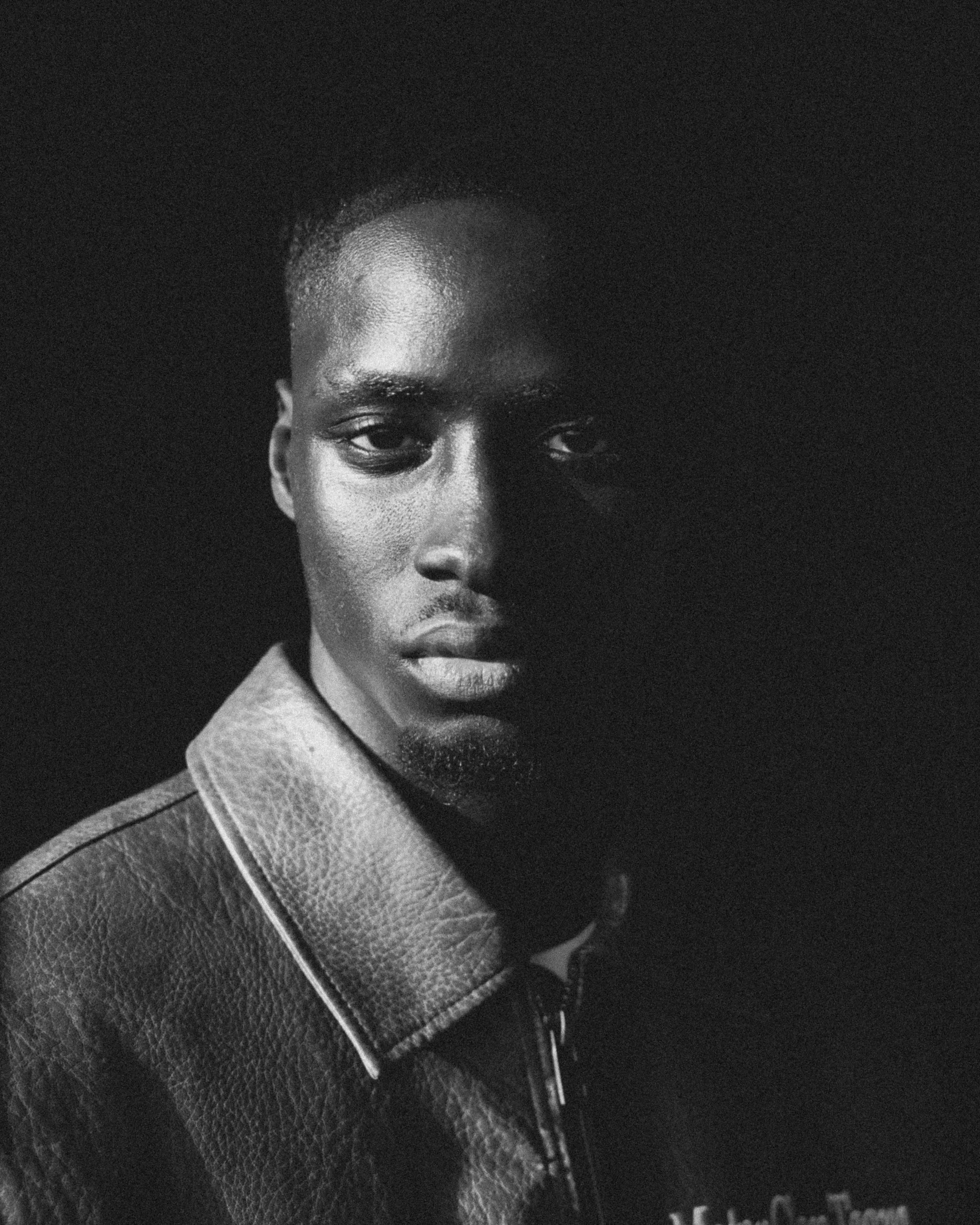 Close-up black-and-white photo of a young person's face with partially visible features, wearing a textured leather jacket and a collared shirt underneath.