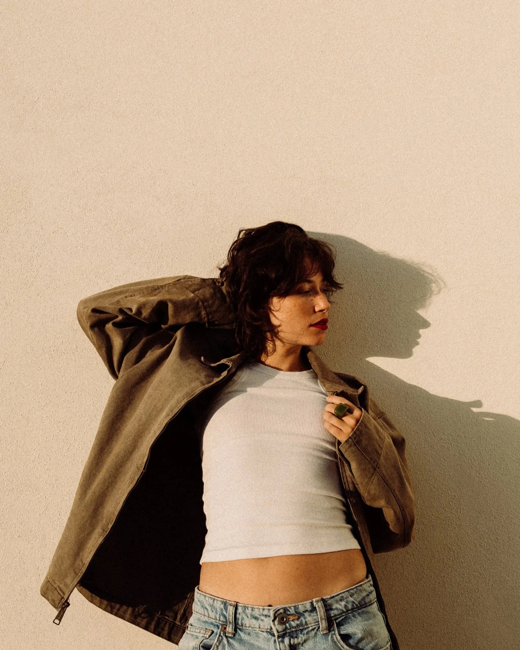 Young woman with dark hair, closed eyes, and red lipstick, wearing a white crop top, denim shorts, and a brown jacket, standing against a beige wall, with her arms raised and hands behind her head.