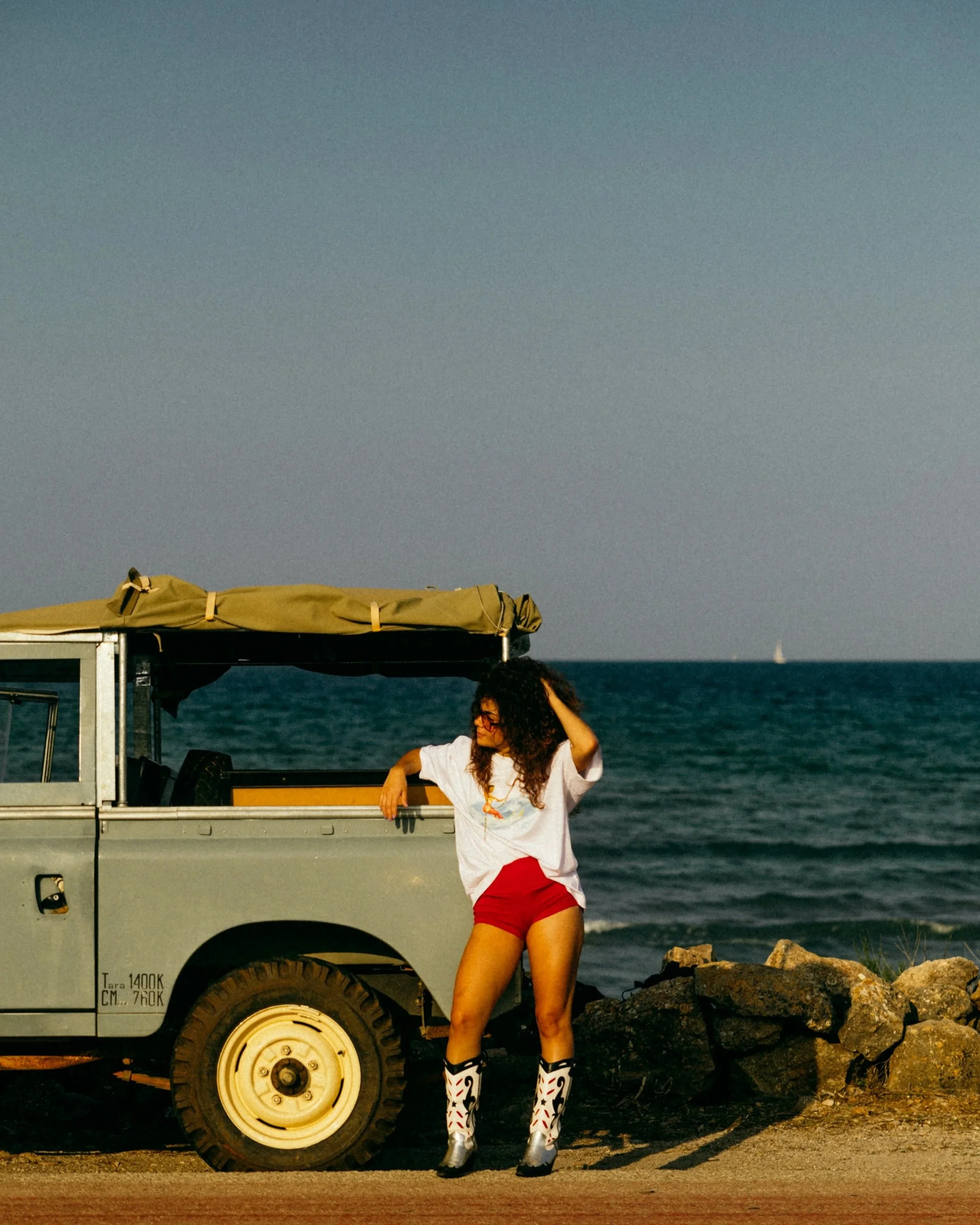A woman with curly hair wearing a white t-shirt, red shorts, and tall patterned boots standing next to a truck by the ocean on a rocky shoreline. The woman appears to be relaxing or posing, and the background features a calm sea and an overcast sky.