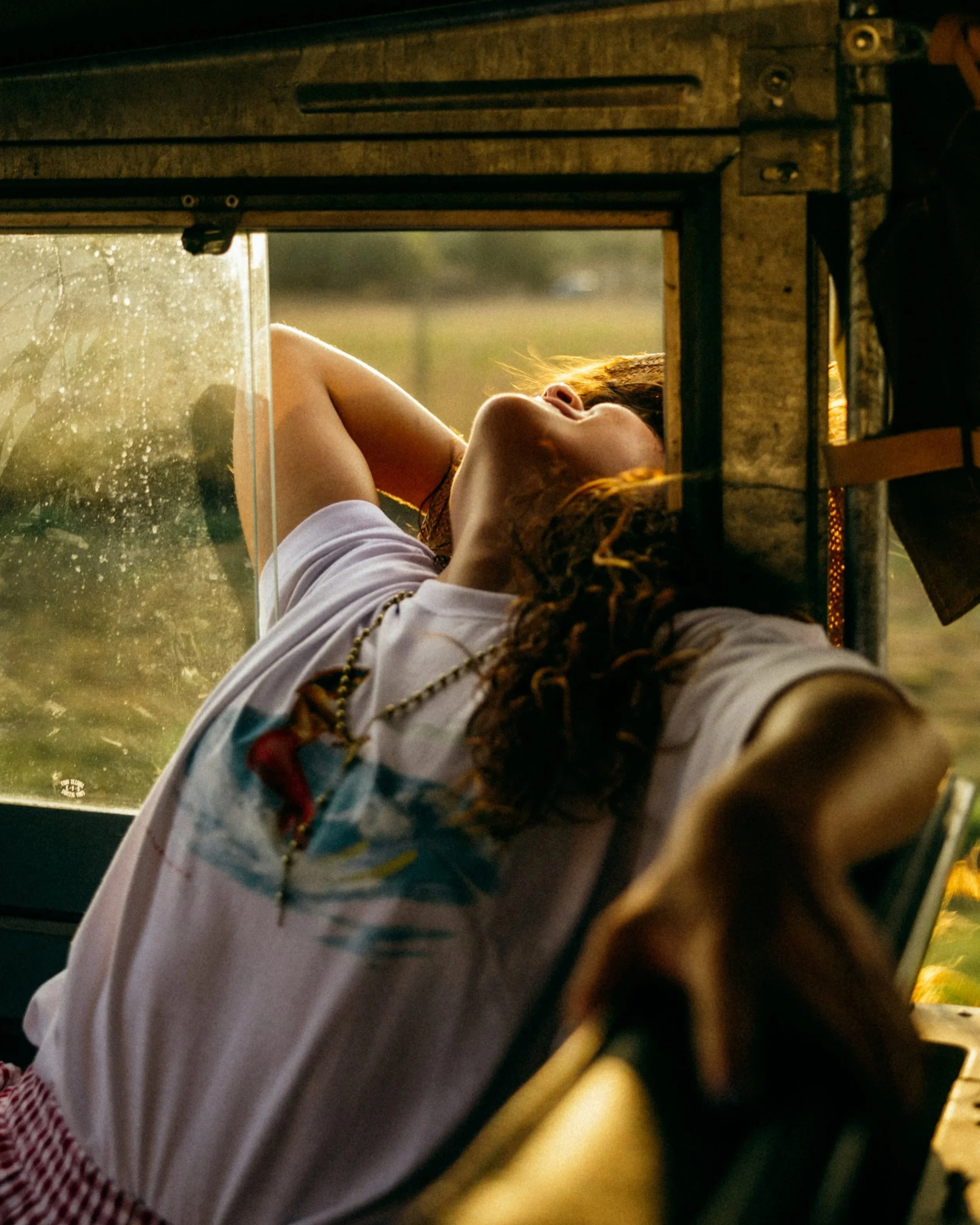 A person with curly hair lying down, visible through a small window with raindrops, at sunset or sunrise. The person is resting and has their arm behind their head.