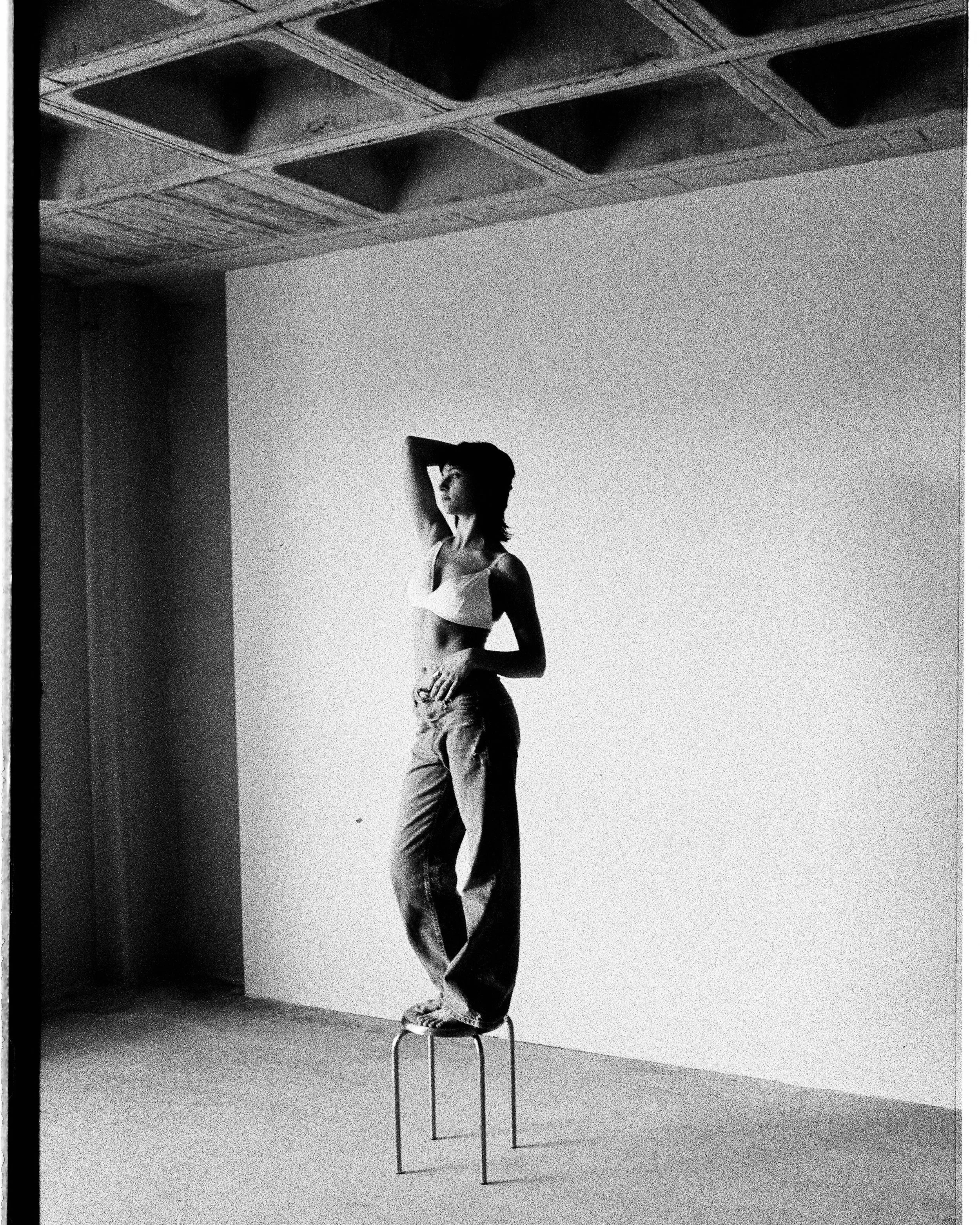 A woman standing on a chair with one hand behind her head and the other on her hip, in a minimalist indoor setting with a plain wall and a ceiling with a grid pattern.