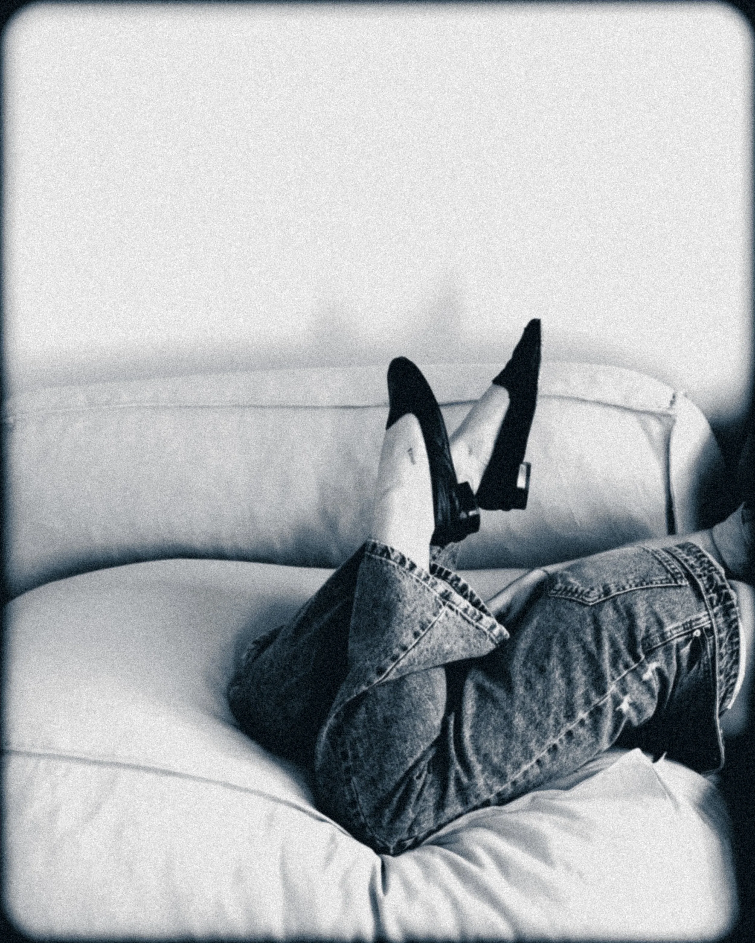 A person wearing jeans and sneakers lying on a bed with white bedding, with their legs crossed in the air, taking a photo from their perspective with a tablet or smartphone.
