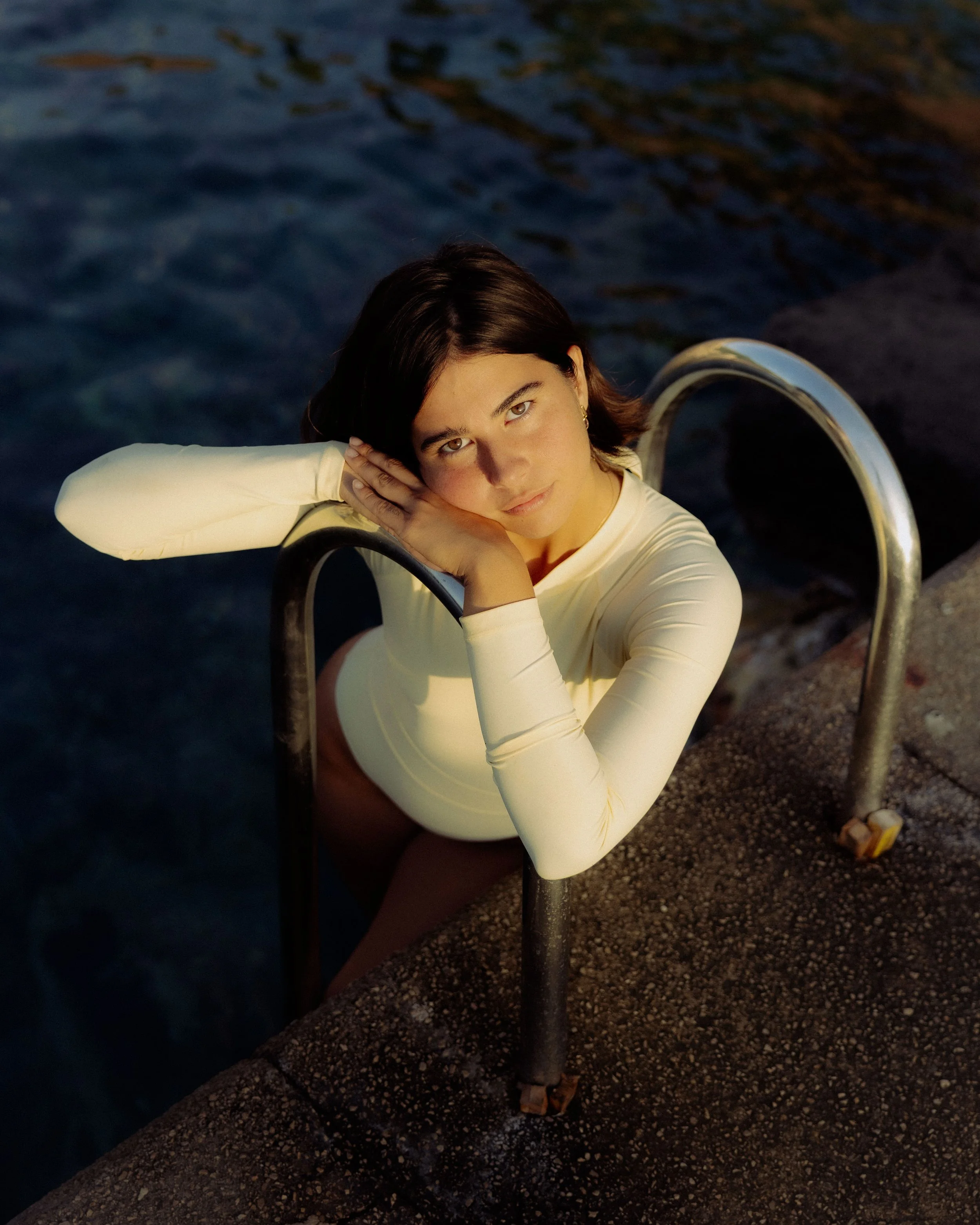 A young woman with short dark hair and fair skin leaning on a ladder by a body of water during sunset.