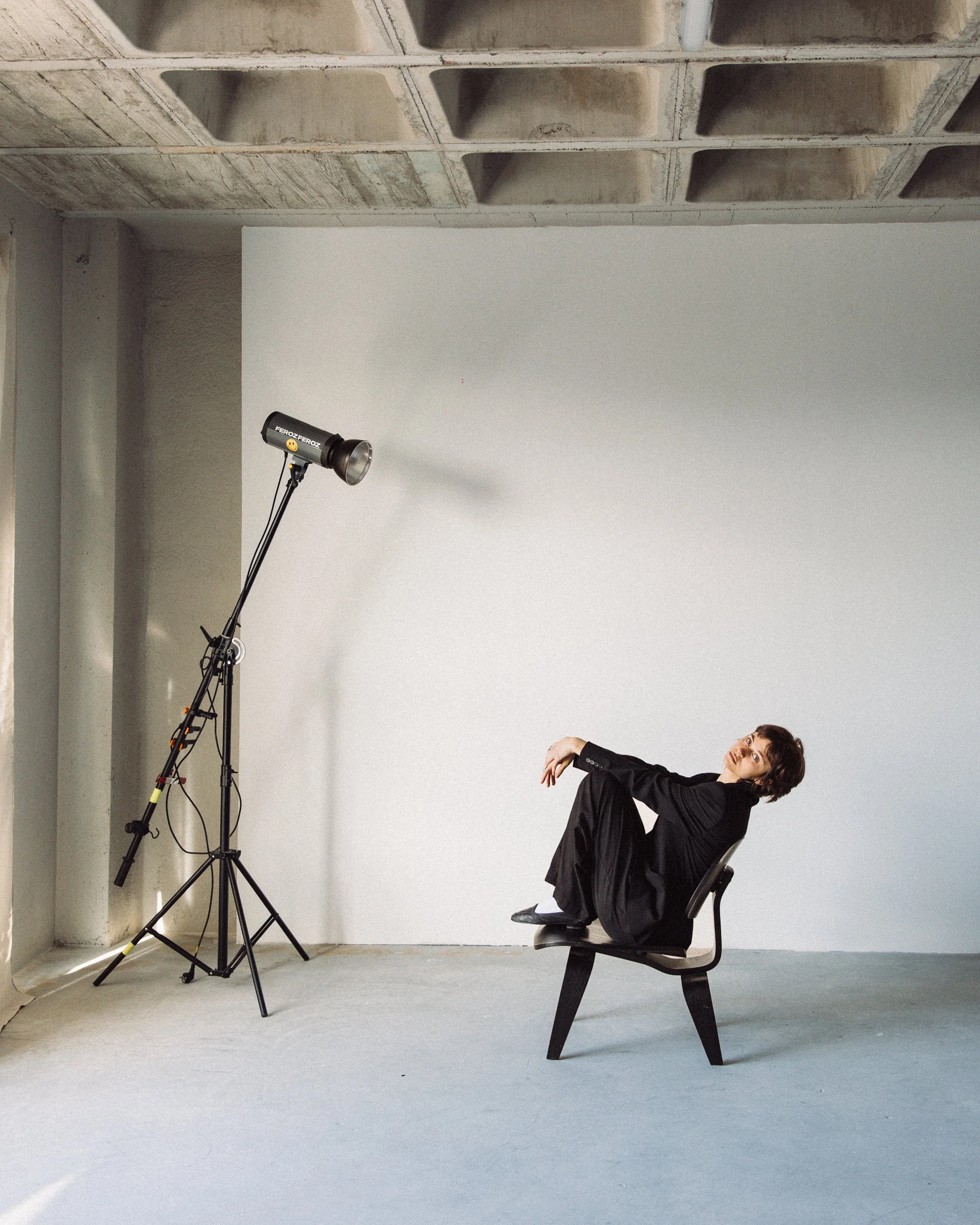 A person with short brown hair, dressed in a black suit, sitting on a chair, leaning back, in a room with a plain white wall and a concrete ceiling with square indentations, next to a tall studio light.