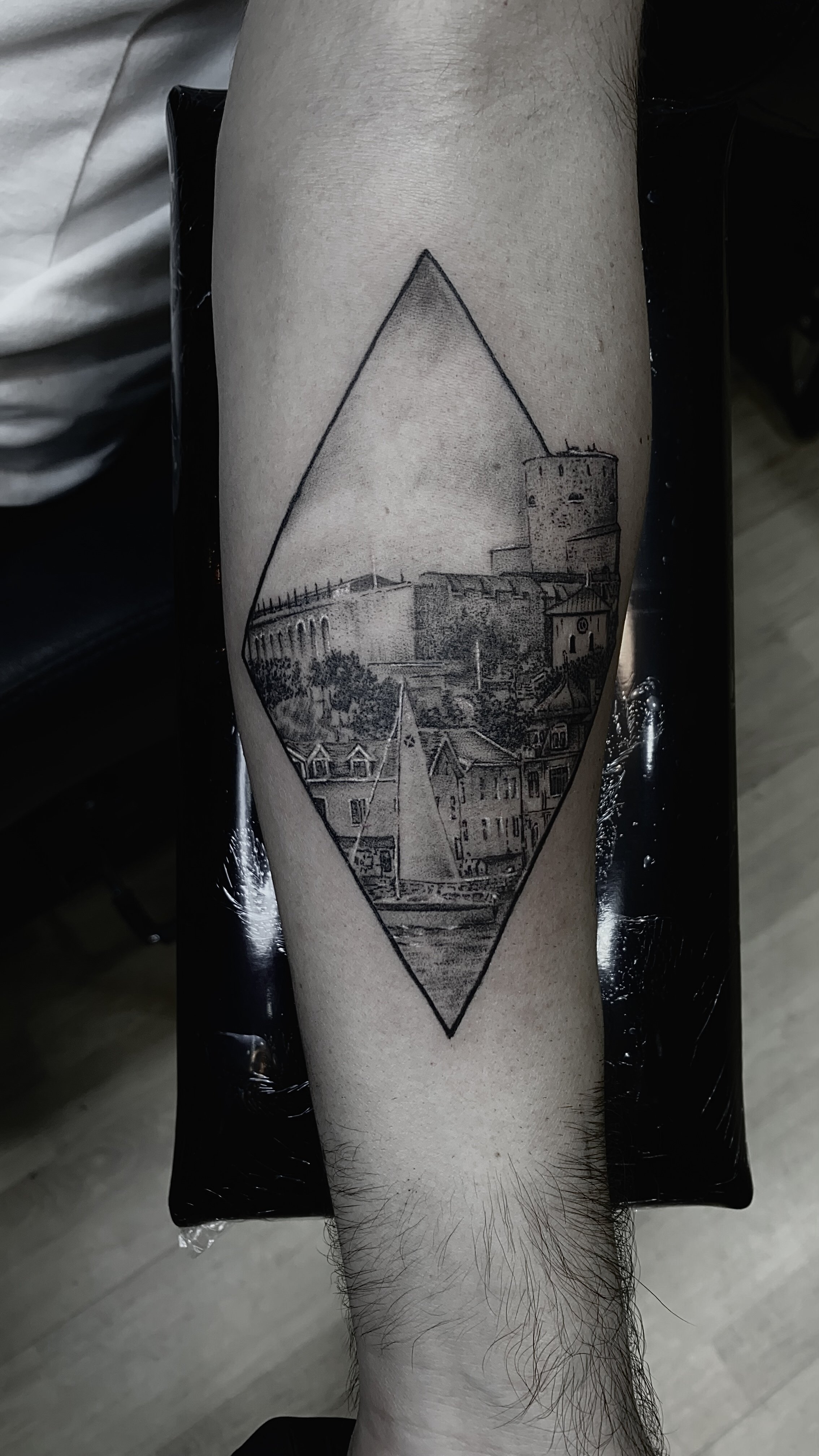 Tatueringsdesign med en segelbåt, byggnader och ett slott inom en diamantform på en mans underben.